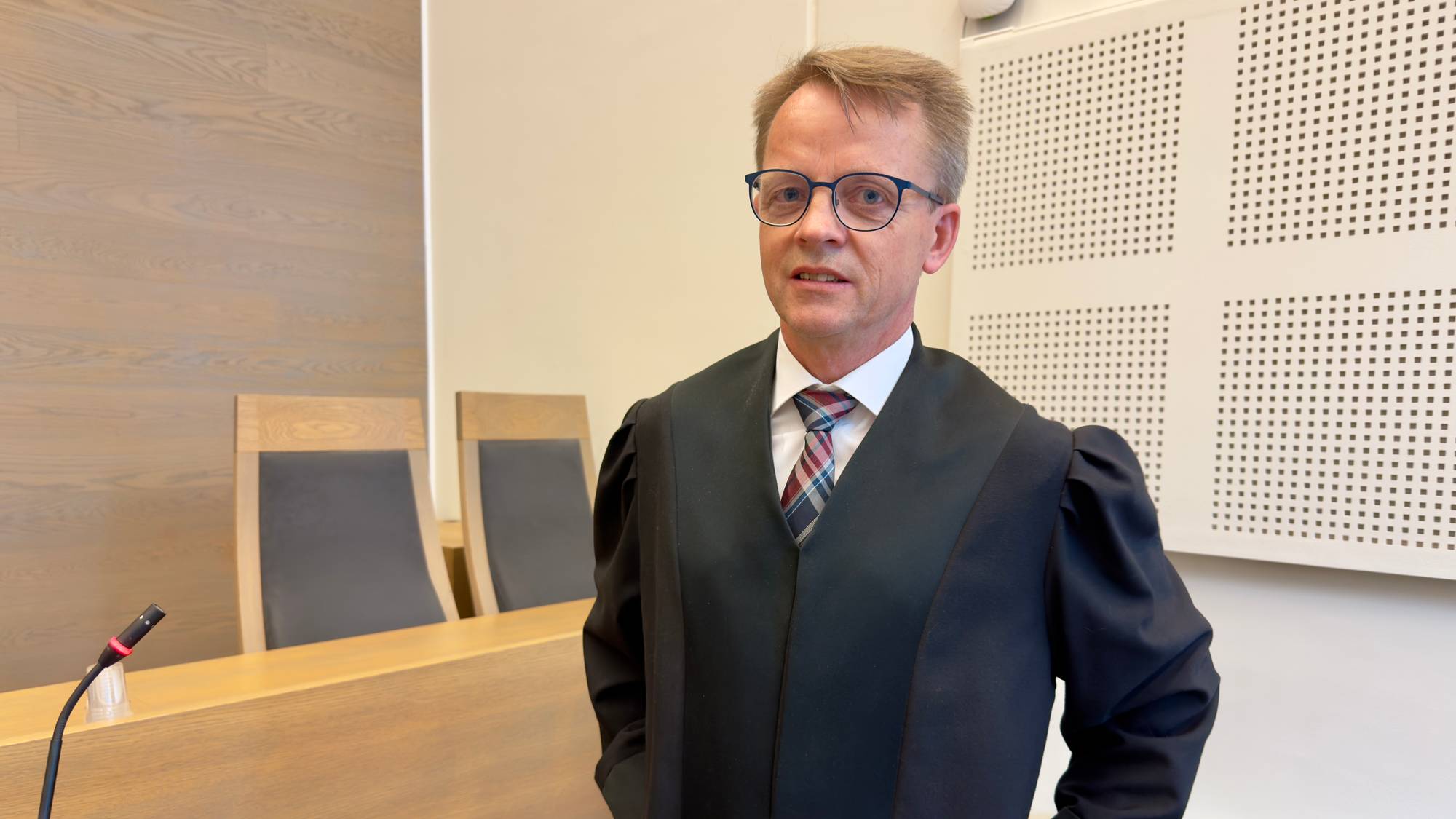 En mann står i sort kappe inne i en rettssal. 