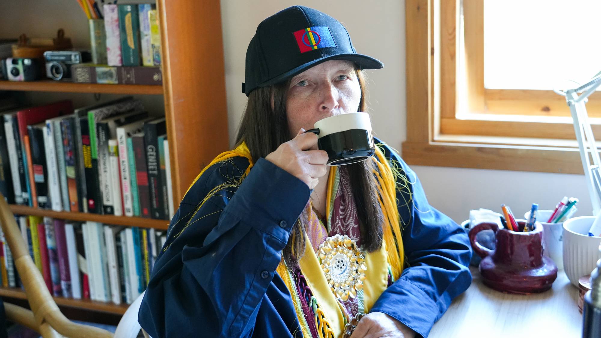 Kvinne med samisk caps og samisk sjal drikker kaffe ved et skrivebord med bokhyller i bakgrunnen. (Bildebeskrivelsen er laget av en KI-tjeneste)