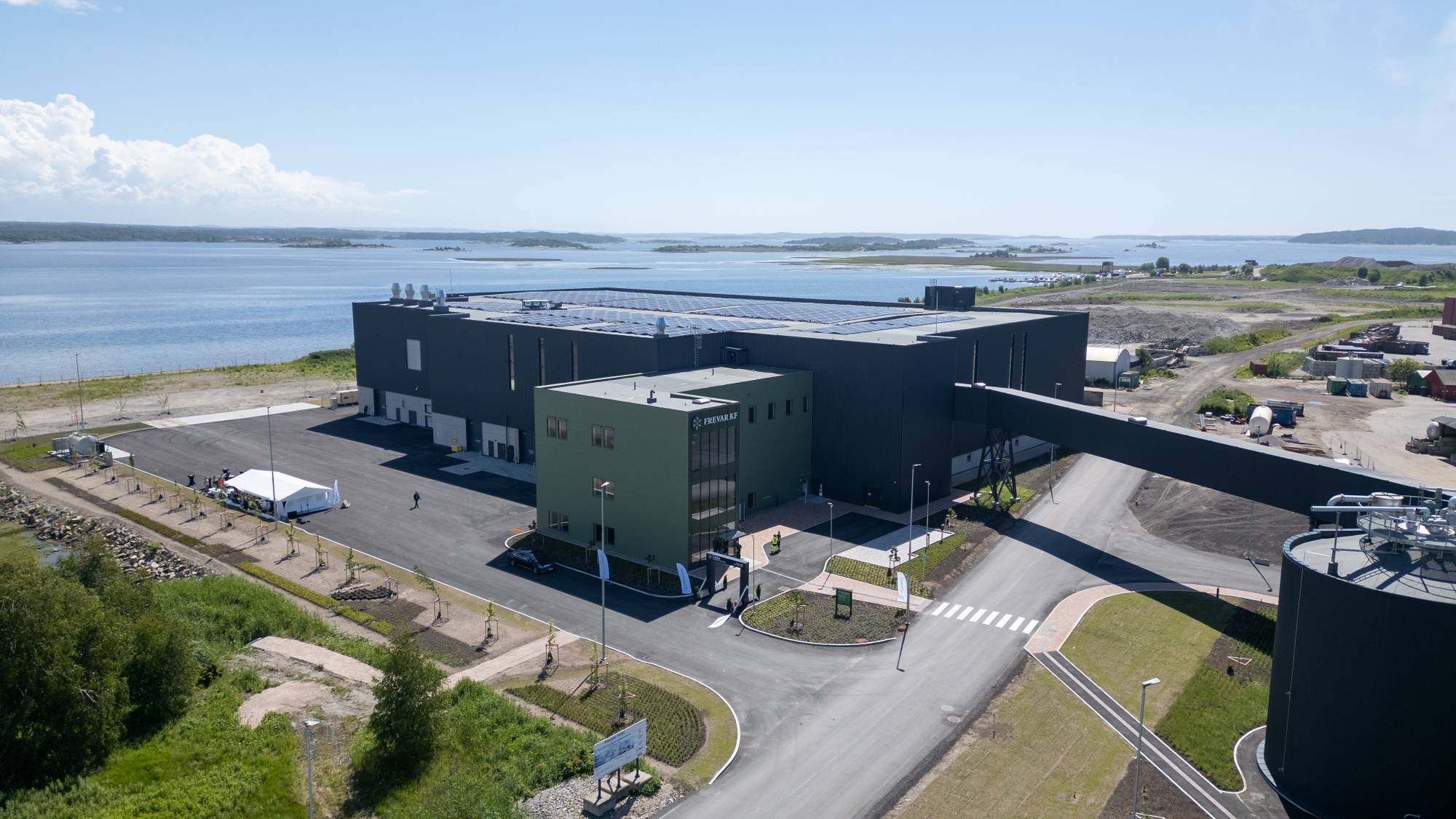 Et nytt industribygg med solcellepaneler på taket er fotografert fra luften. Bygget har et tilknyttet transportbånd og en stor lagertank. En hvit presenning er satt opp ved en inngang til bygget. Flere biler og noen personer er synlige på området. (Bildebeskrivelsen er laget av en KI-tjeneste)