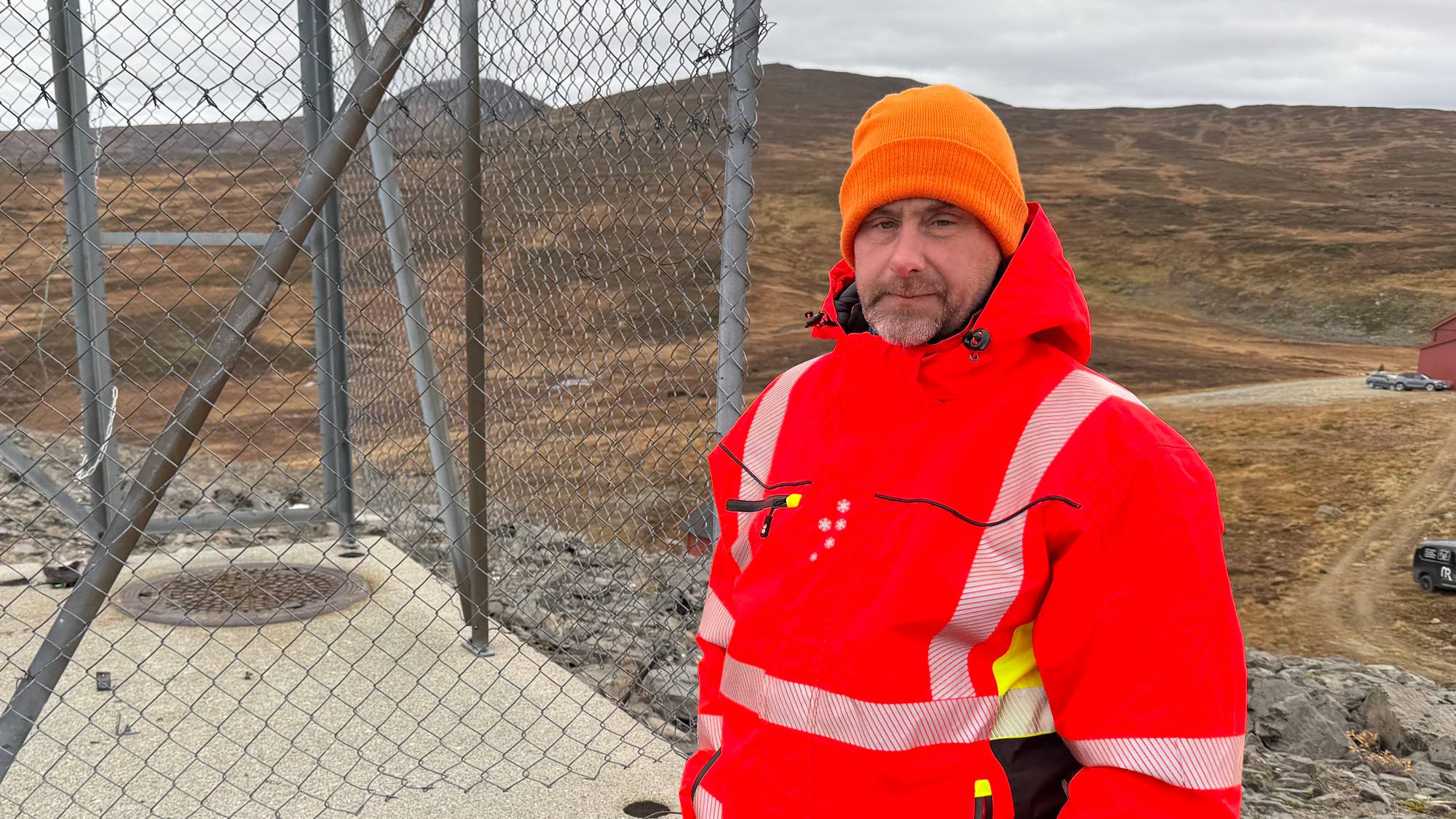 Mann i oransje vernetøy og lue står ved et inngjerdet teknisk anlegg i et fjellandskap. (Bildebeskrivelsen er laget av en KI-tjeneste)