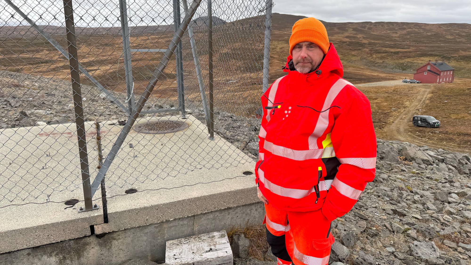 Mann i oransje vernetøy og lue står ved et inngjerdet teknisk anlegg i et fjellandskap. (Bildebeskrivelsen er laget av en KI-tjeneste)