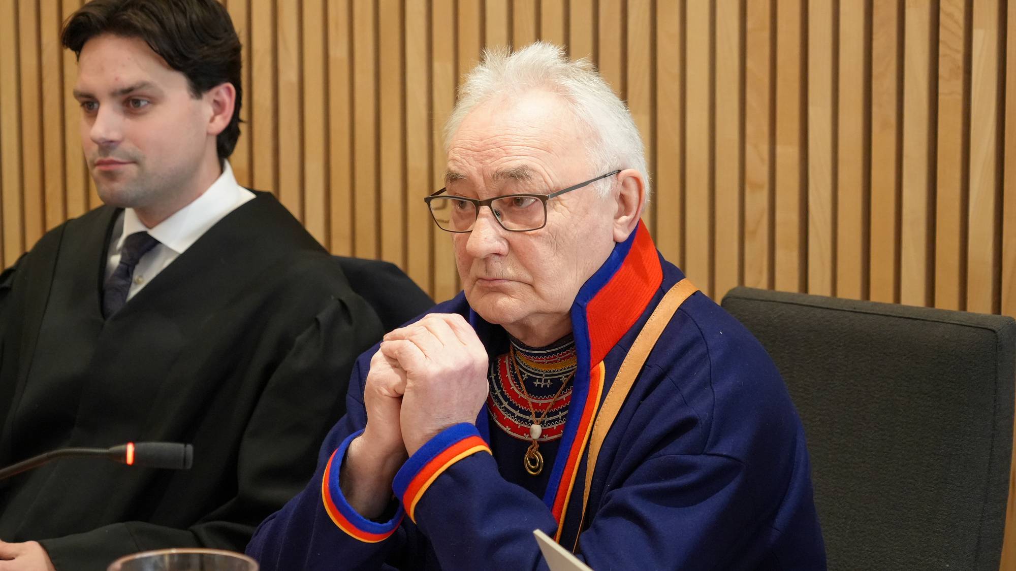 en mann som sitter i en stol med en mann stående bak seg (Bildebeskrivelsen er laget av en KI-tjeneste)