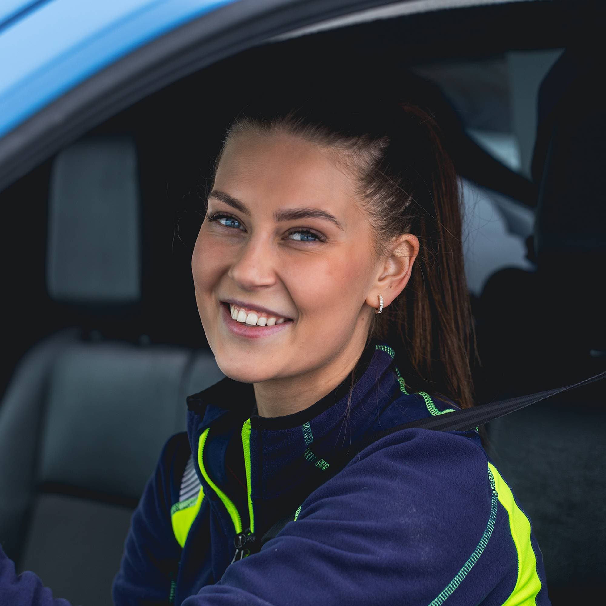 Lea sitter bak rattet i bilen sin, hun ser i kamera og smiler pent, de blå øynene hennes lyser opp av snøen utenfor bilen.