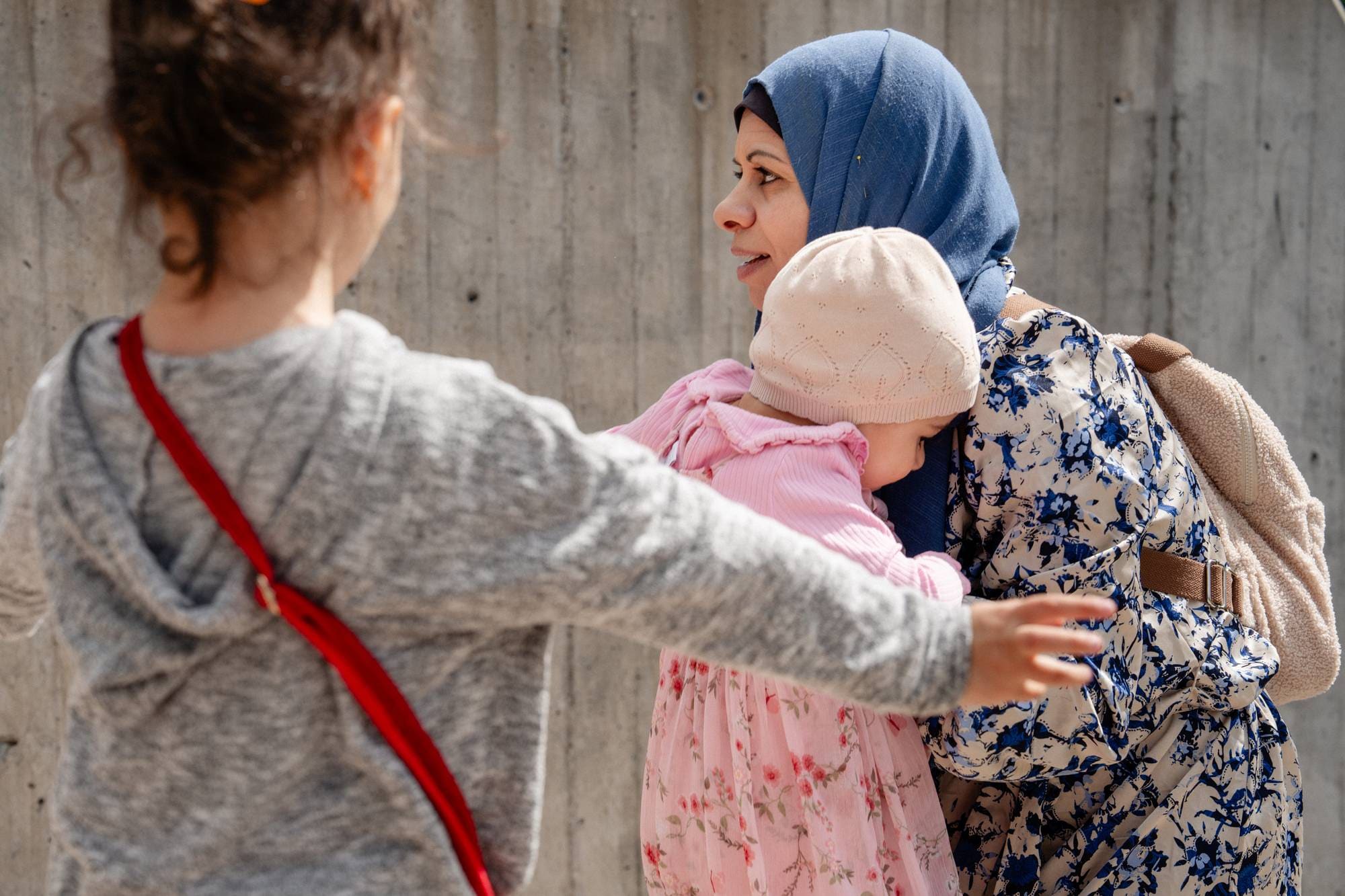 En kvinne med hijab klemmer et lite barn med rosa kjole og lue. Et annet eldre barn står i forgrunnen og ser på. 
