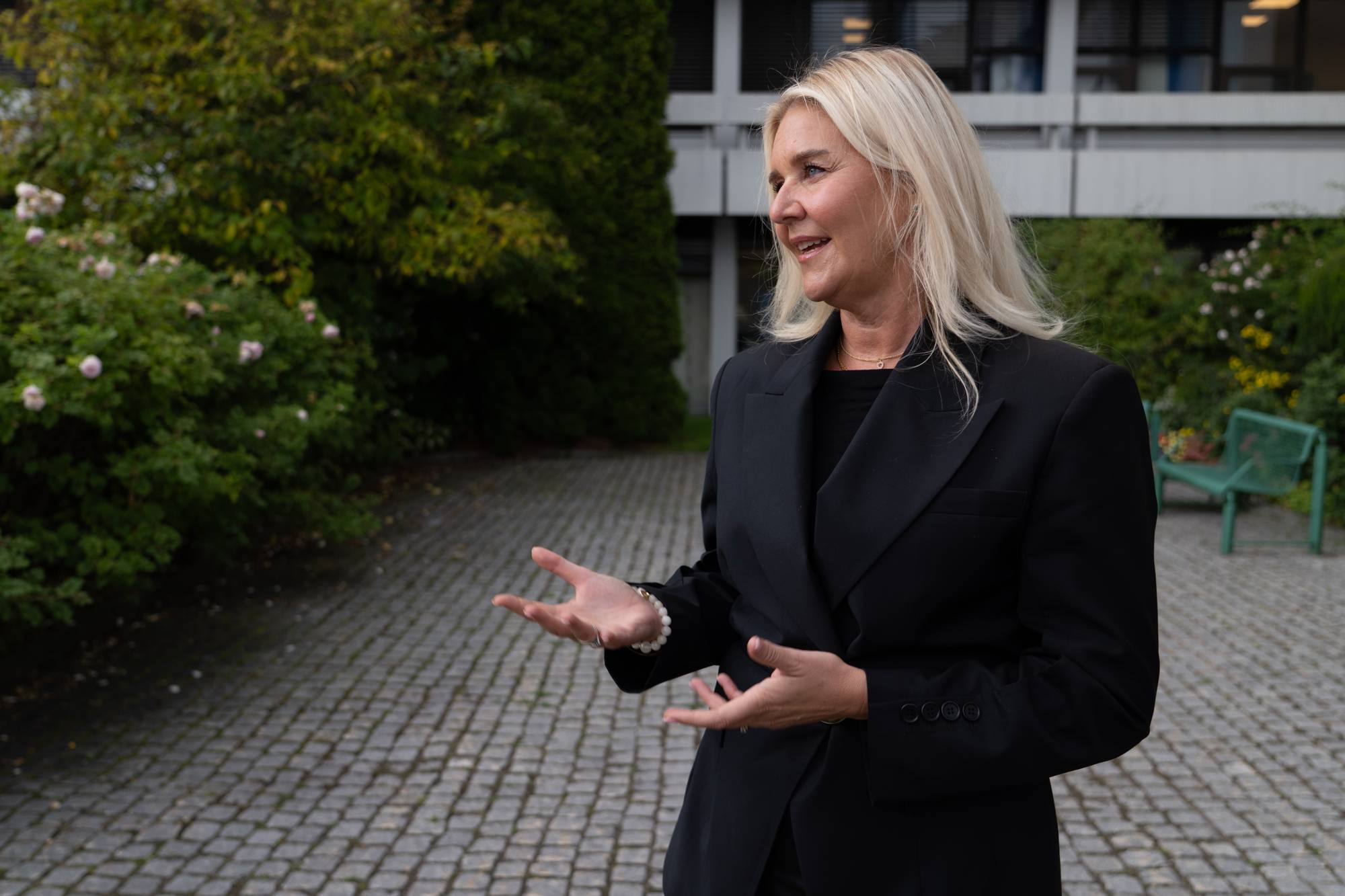Kvinne med blondt hår i svart dress gestikulerer utendørs foran en bygning. (Bildebeskrivelsen er laget av en KI-tjeneste)