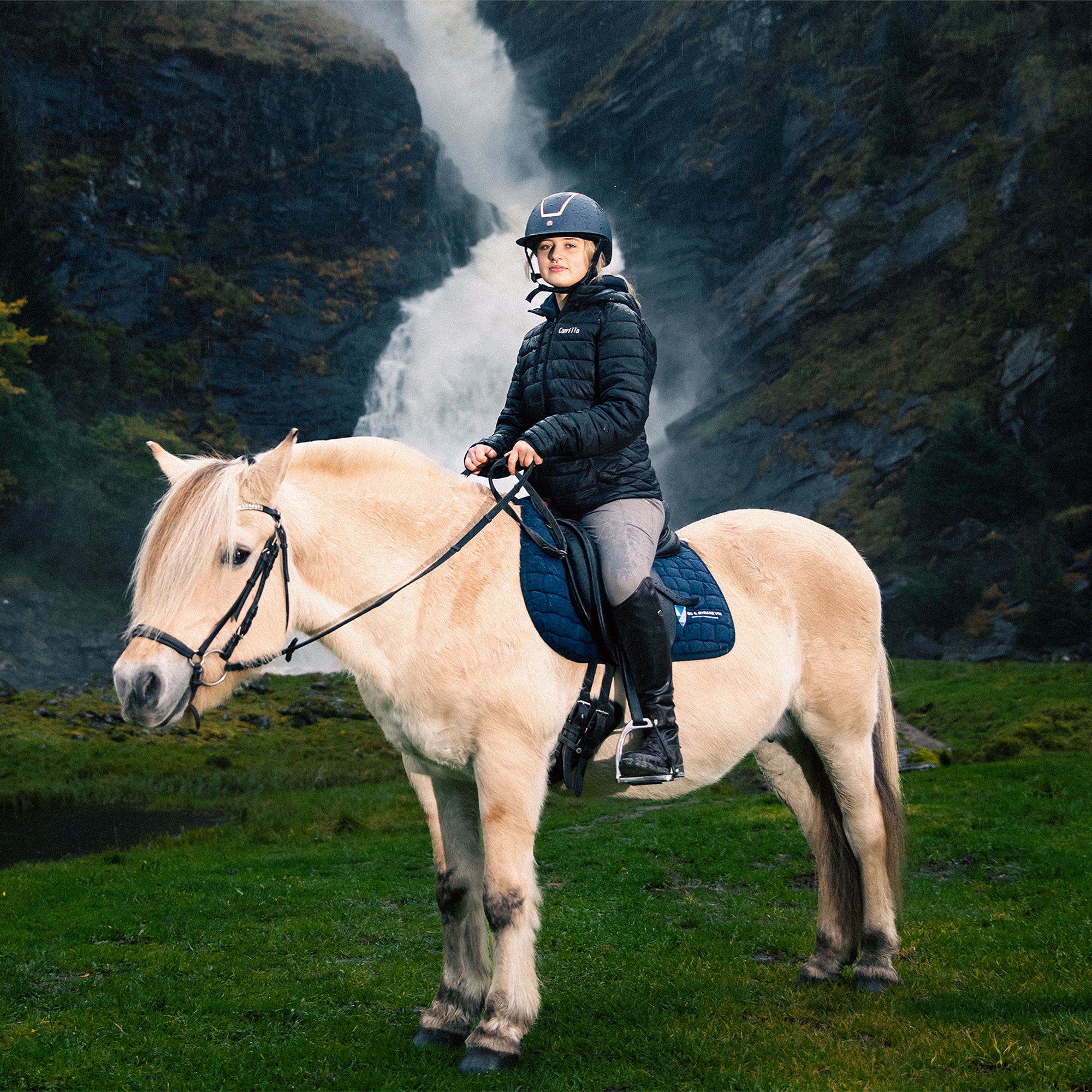 Bilde av skribenten, Camilla, på en hest. I bakgrunnen ser du en stor foss i et frodig landskap. 