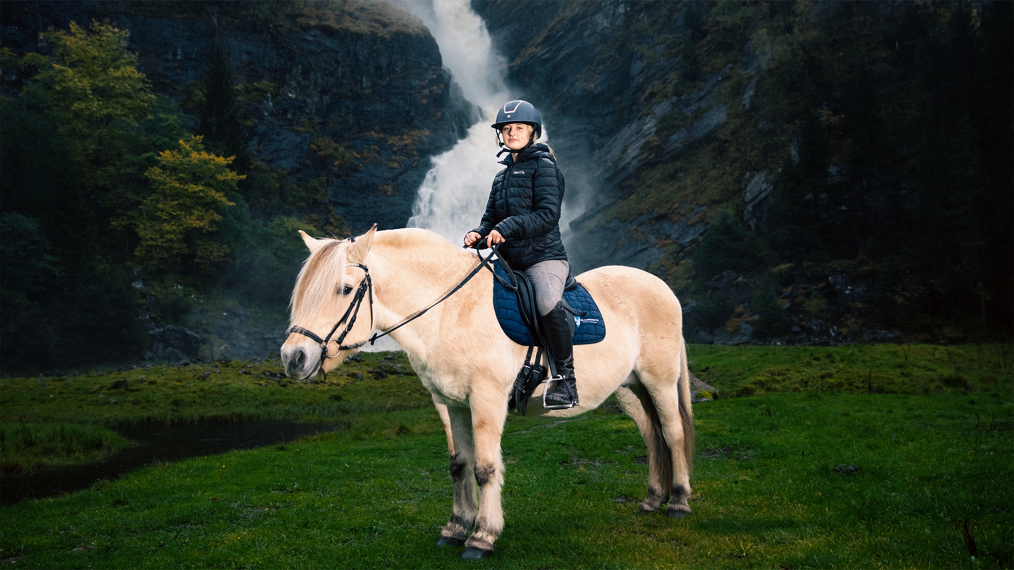 Bilde av skribenten, Camilla, på en hest. I bakgrunnen ser du en stor foss i et frodig landskap. 