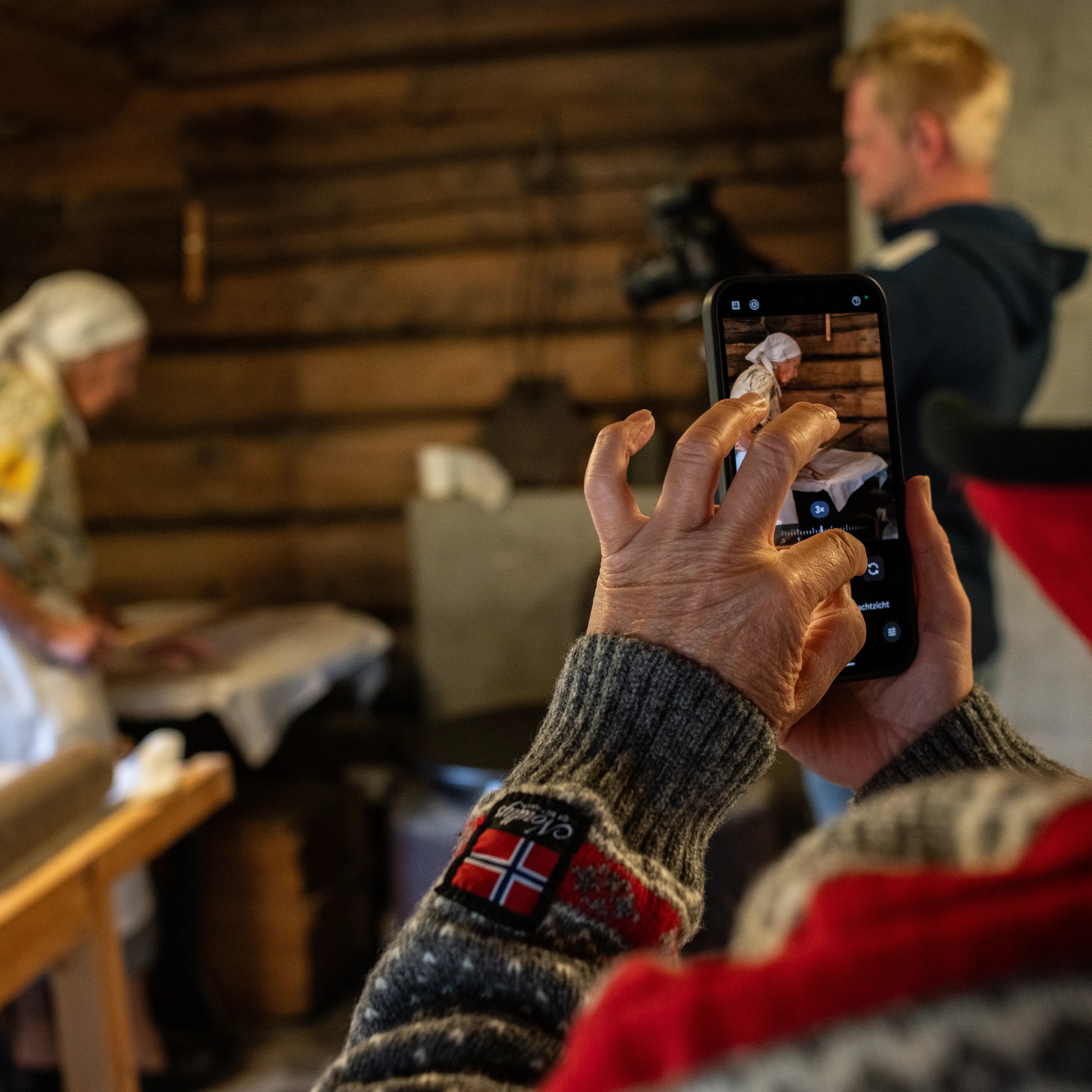 Person fotograferer kvinne som baker i et tømmerhus med mobiltelefon.