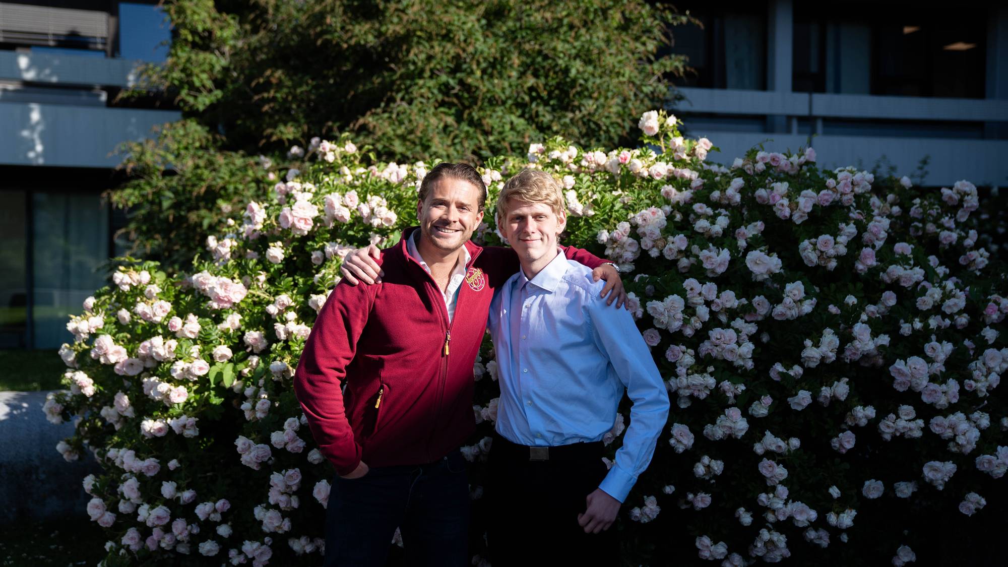 To menn står med armene rundt hverandre foran en rosebusk. Den ene mannen har på seg en rød fleecegenser og mørke bukser. Den andre mannen har på seg en lyseblå skjorte og mørke bukser. I bakgrunnen er det en bygning og trær. (Bildebeskrivelsen er laget av en KI-tjeneste)