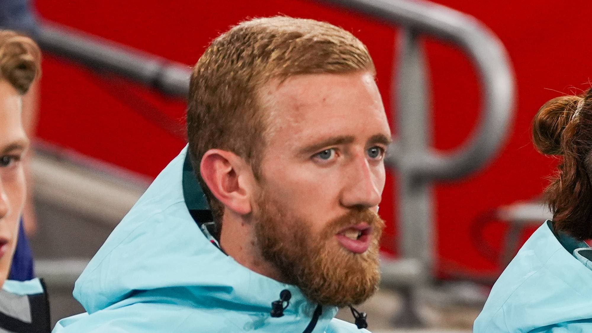 Norske landslagsspillere i lyseblå treningsjakker sitter på benken under en fotballkamp. (Bildebeskrivelsen er laget av en KI-tjeneste)