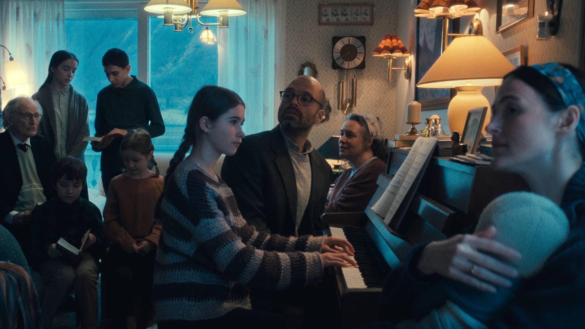 En gruppe mennesker står og sitter i en stue. En jente spiller piano.