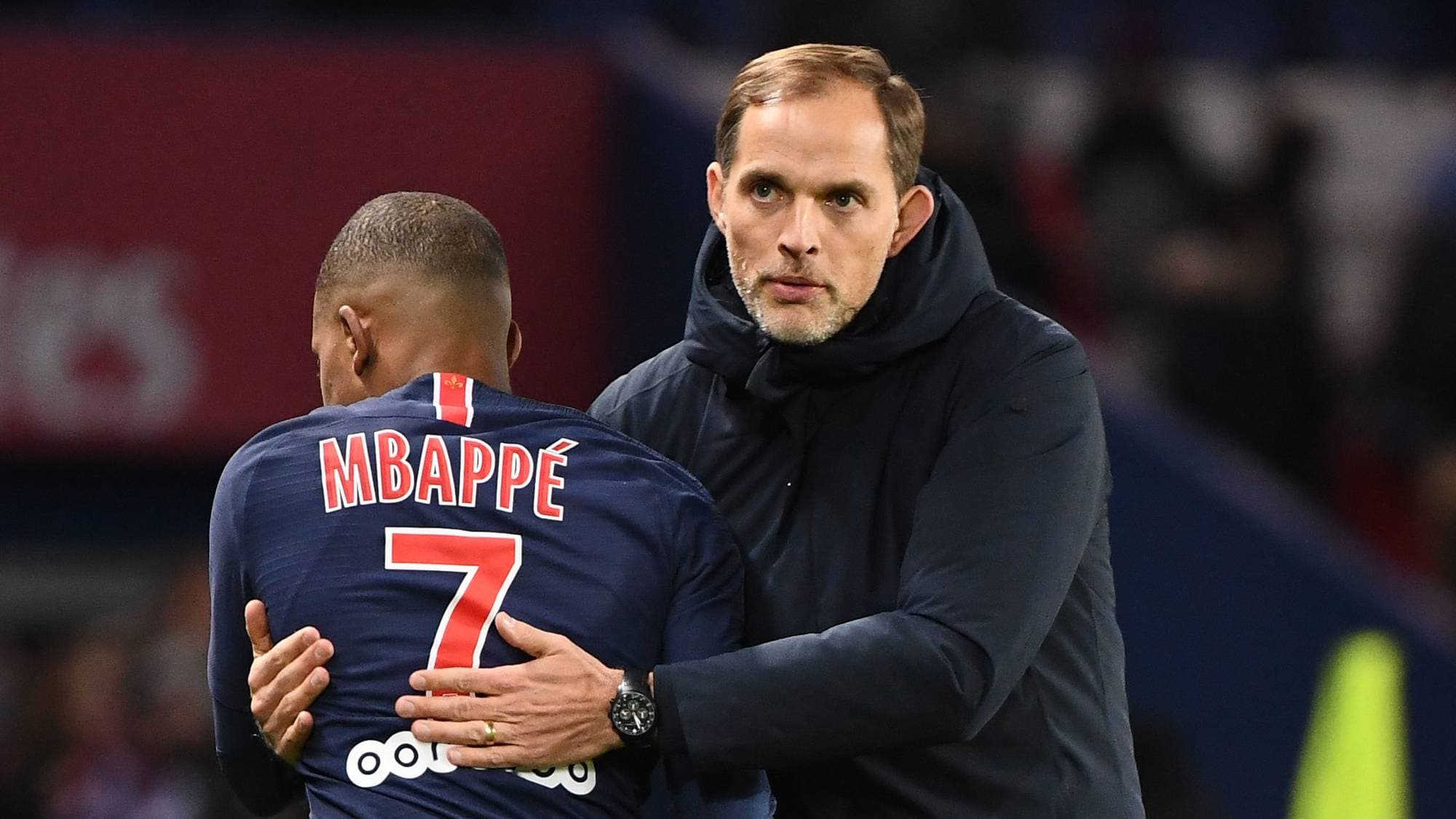 Fotballtrener Thomas Tuchel legger armen rundt Kylian Mbappé på en fotballbane. (Bildebeskrivelsen er laget av en KI-tjeneste)