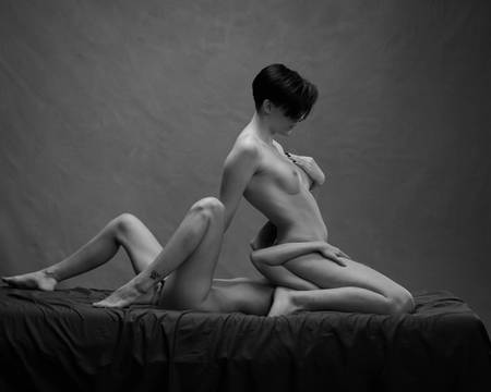 En kvinne med kort mørkt hår kneler over en annen kvinne med blondt hår som ligger på ryggen. Hun sitter over ansiktet hennes med beina spredt. De simulerer oralsex. Begge er nakne, men kjønnsorganene er ikke synlige.
