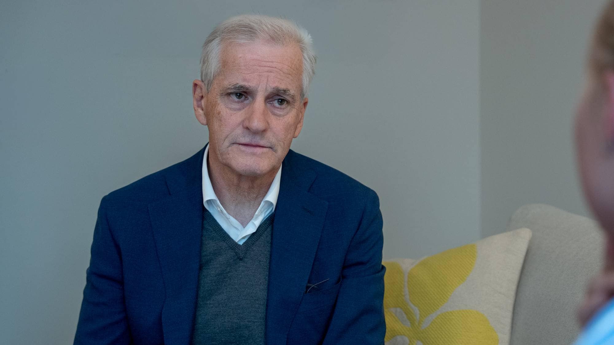 En mann i dress sitter ved siden av en person som er ute av fokus. Mannen har grått hår og ser mot personen. De sitter i et rom med en grå vegg og en sofa. Det er en pute med gult mønster på sofaen. (Bildebeskrivelsen er laget av en KI-tjeneste)