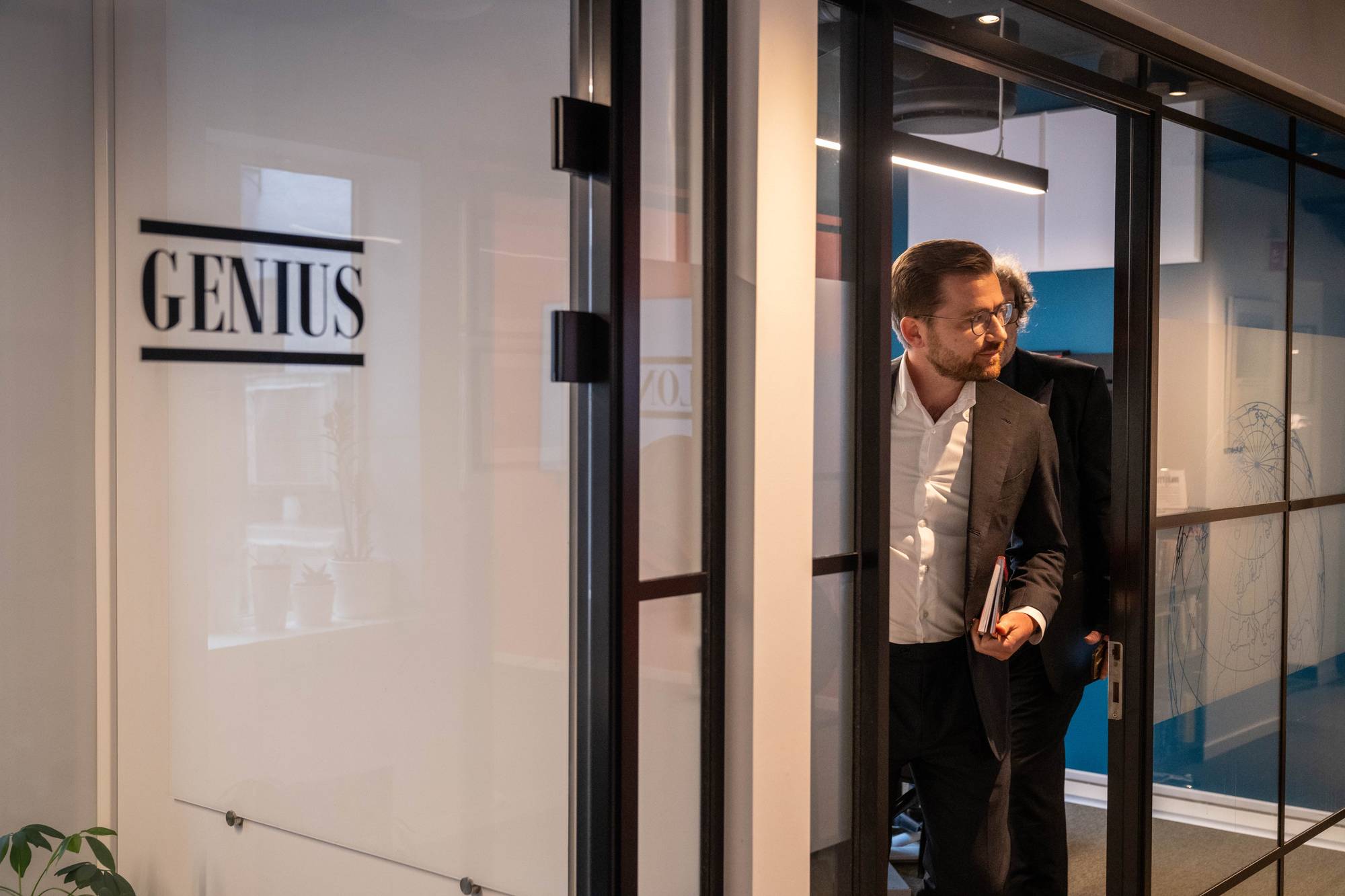 En mann med briller og dress går gjennom en glassdør. Ved siden av ham går en annen person i dress. Et skilt med teksten "GENIUS" er synlig på glassveggen. Inne i rommet ses moderne belysning og et kart på en glassvegg. (Bildebeskrivelsen er laget av en KI-tjeneste)