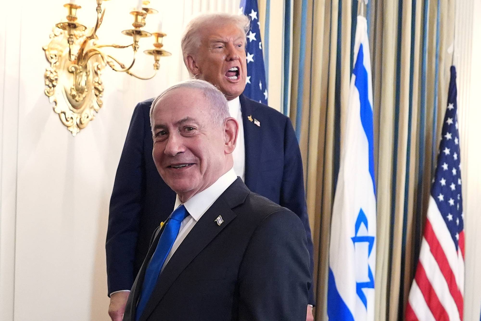 Benjamin Netanyahu i dress smiler, bak ham står Donald Trump og ser ut som om han roper. I bakgrunnen det israelske og det amerikanske flagget.