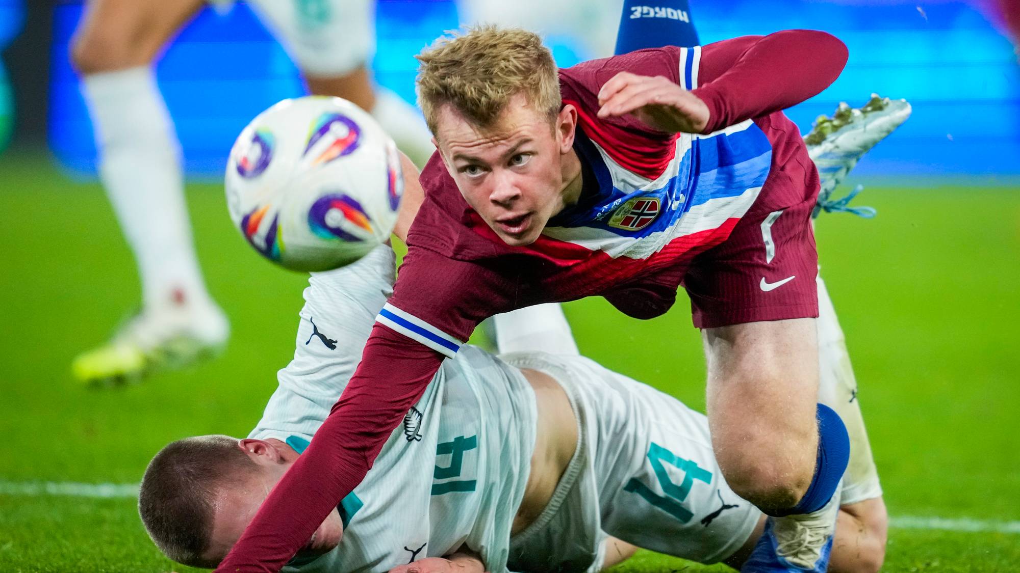 En spiller iført rød og blå drakt faller over en spiller med hvit drakt på en fotballbane. Begge spillerne er nær bakken, med en ball i luften. Omgivelsene viser en stadion med lyseffekter i bakgrunnen. Det er utydelig hvor kampen pågår.
