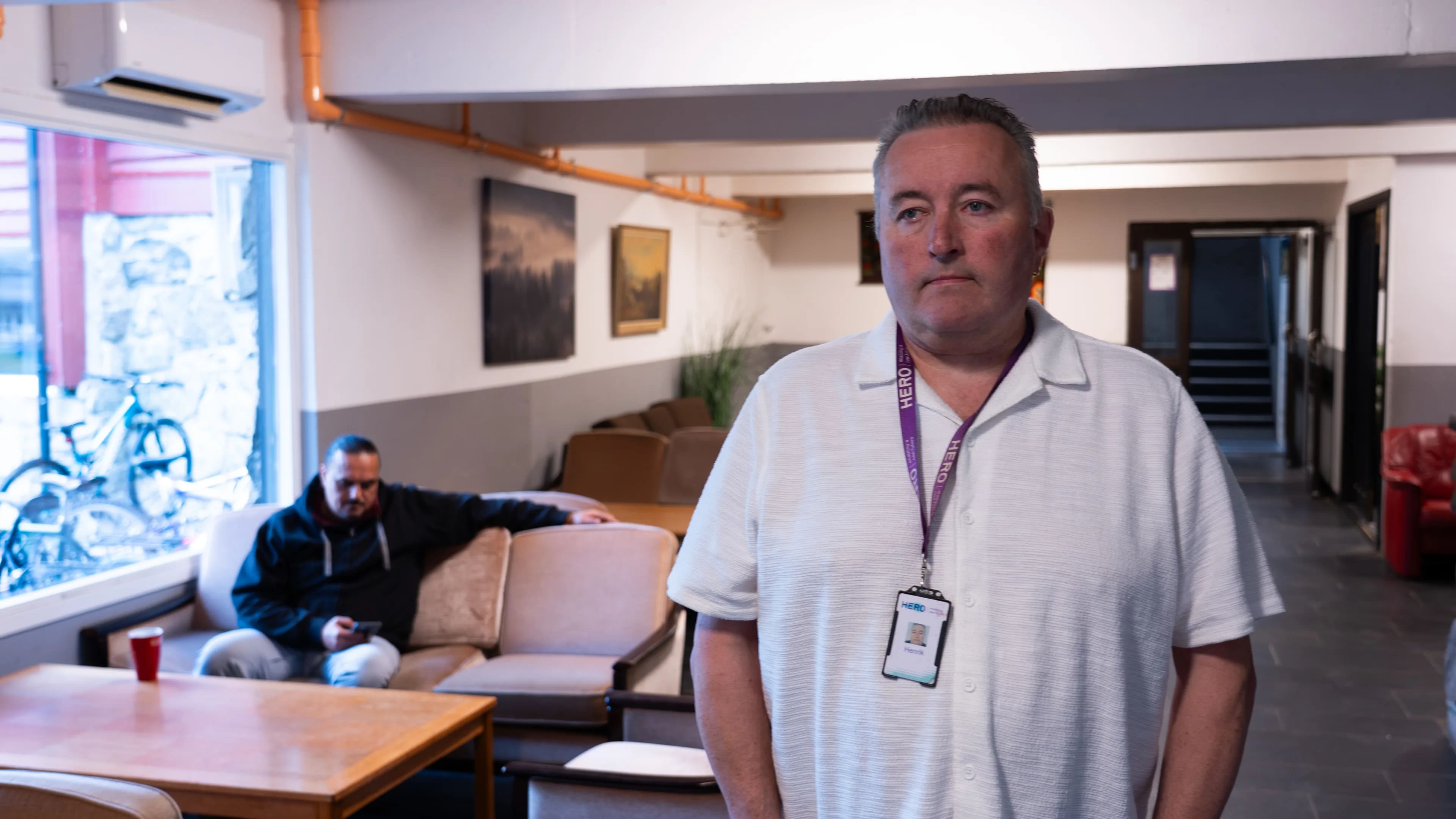 Mann med adgangskort i et fellesrom. En annen person sitter i bakgrunnen. (Bildebeskrivelsen er laget av en KI-tjeneste)