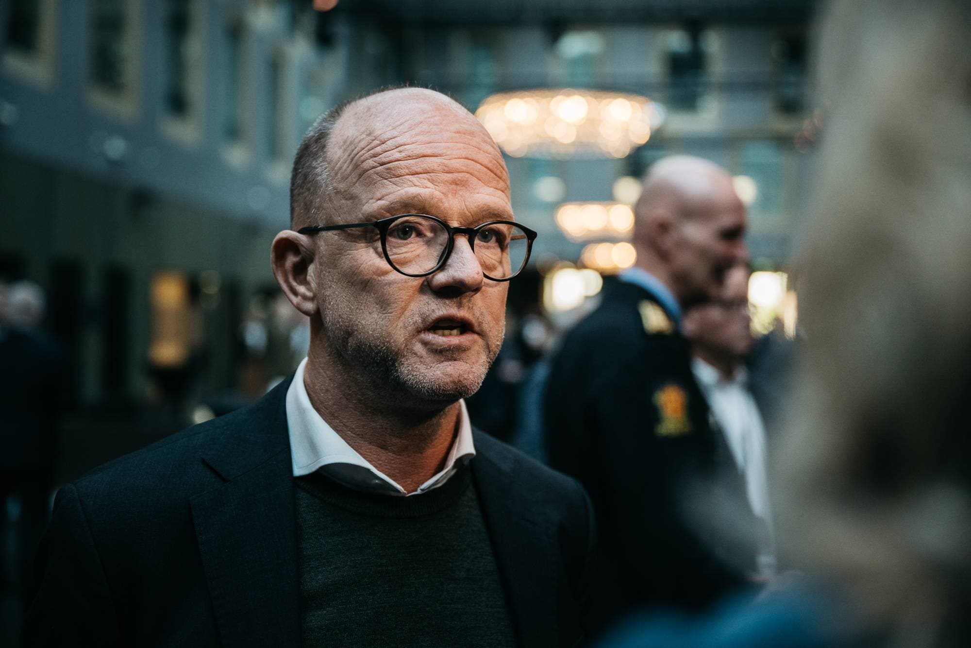 Bildet viser en gruppe mennesker i en innendørssetting, delvis omkranset av fargerike lamper. To menn i forgrunnen står nær hverandre, mens flere andre personer er synlige i bakgrunnen. Den ene mannen er kledd i en mørk dress, mens den andre har en uniformert antrekk. Atmosfæren ser ut til å være formell, muligens relatert til en konferanse eller et møtested.