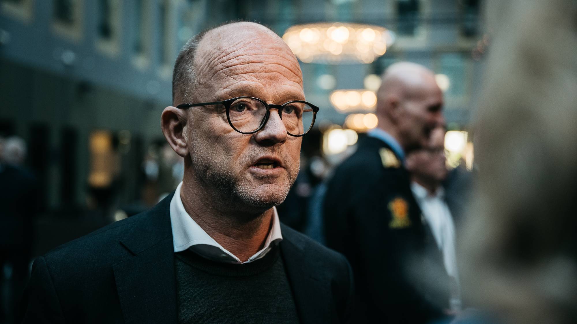 Bildet viser en gruppe mennesker i en innendørssetting, delvis omkranset av fargerike lamper. To menn i forgrunnen står nær hverandre, mens flere andre personer er synlige i bakgrunnen. Den ene mannen er kledd i en mørk dress, mens den andre har en uniformert antrekk. Atmosfæren ser ut til å være formell, muligens relatert til en konferanse eller et møtested.