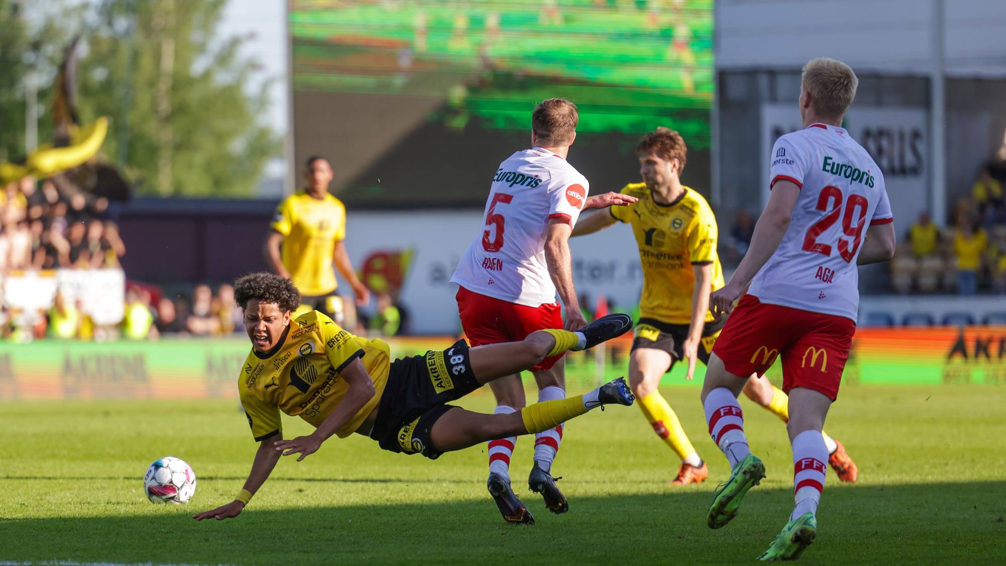 En fotballspiller i gult faller mot bakken. En annen spiller i hvitt forsøker å takle ham. Flere spillere er synlige i bakgrunnen på fotballbanen. (Bildebeskrivelsen er laget av en KI-tjeneste)
