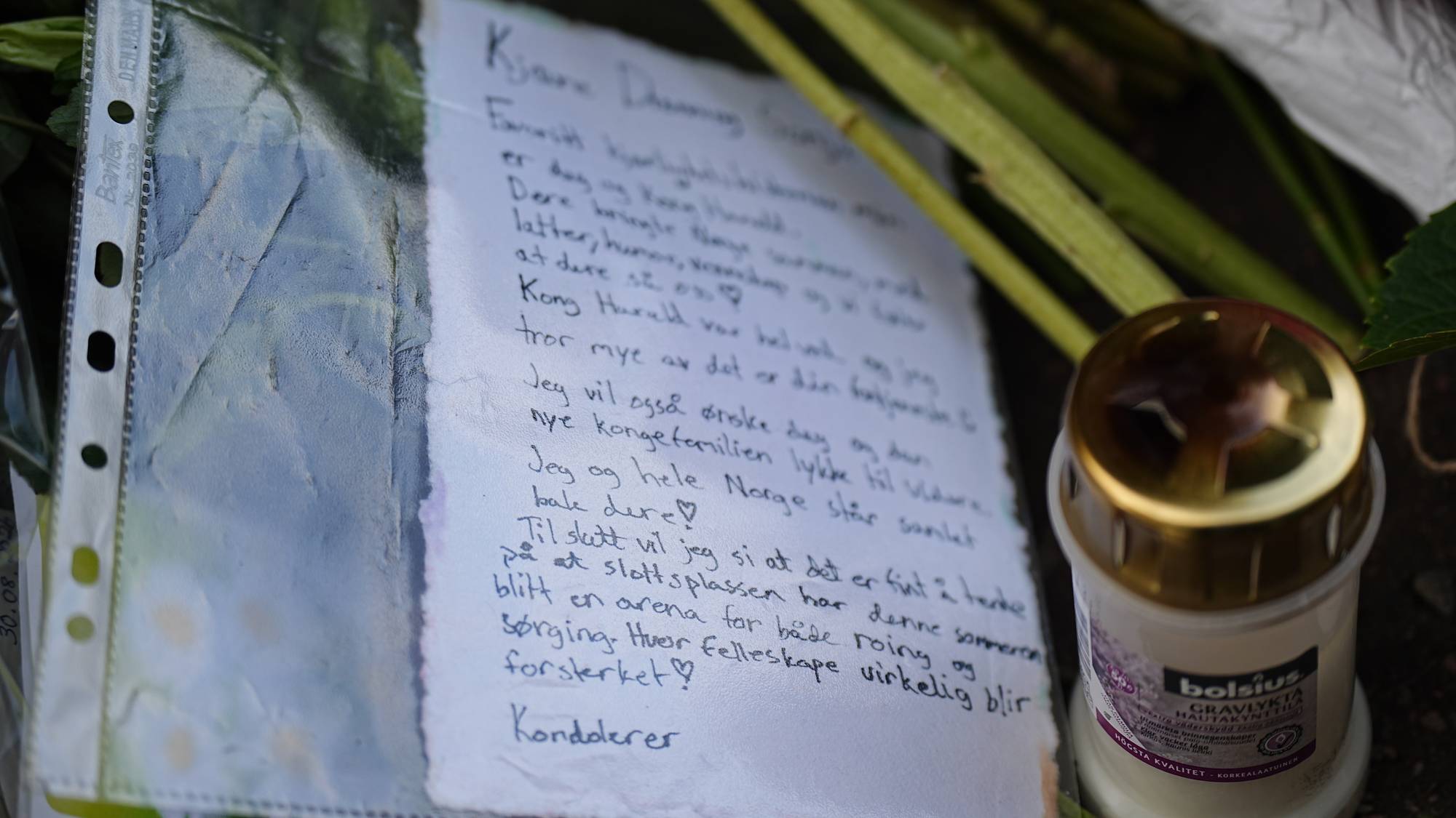 Håndskrevet hilsen til kongefamilien plassert ved blomster og et gravlys. (Bildebeskrivelsen er laget av en KI-tjeneste)