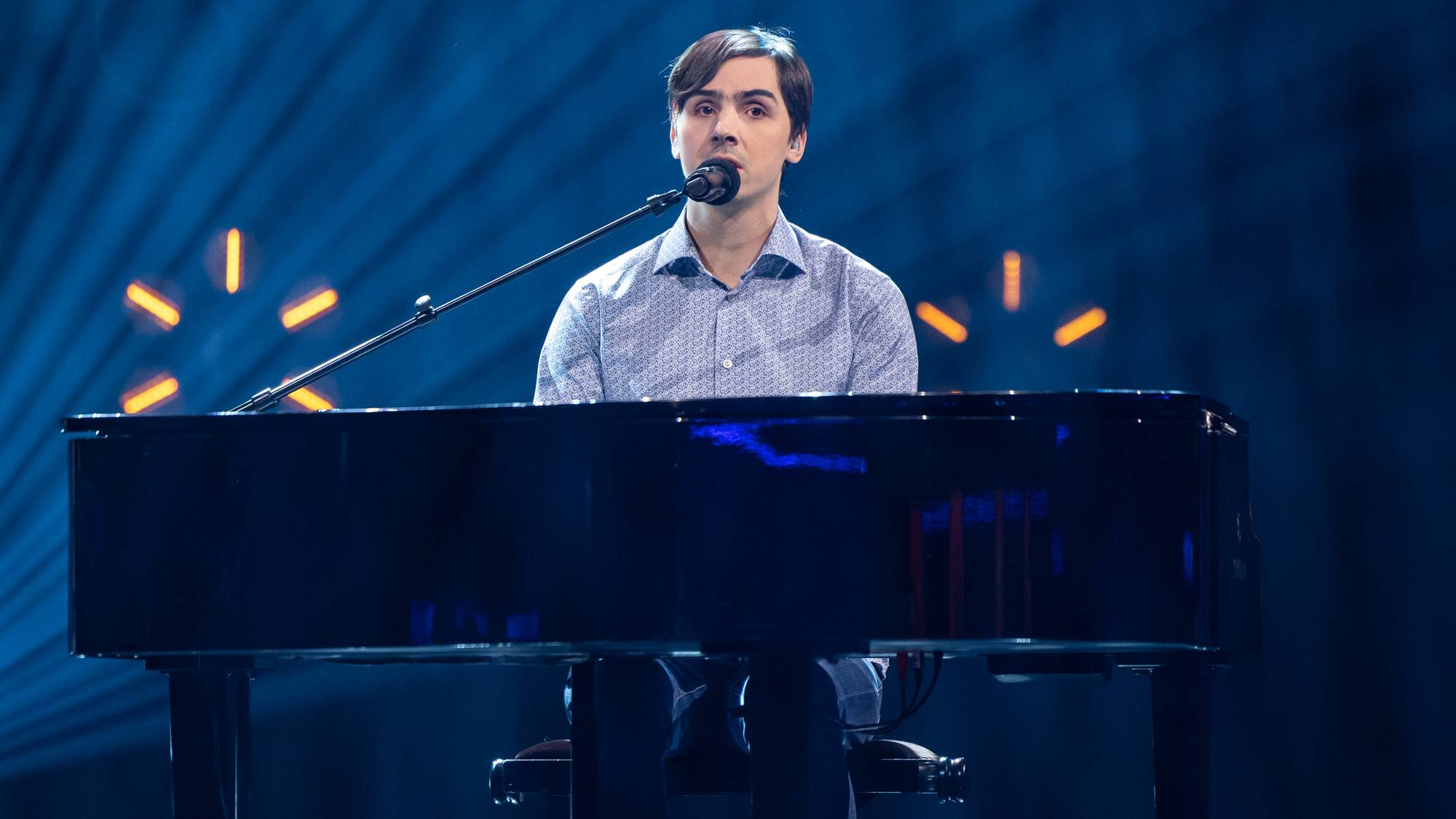 En mann i lysblå skjorte sitter ved et piano og synger, han sitter på scenen og har en lang mikrofon foran seg som stikker opp bak pianoet. Bak ham er lys i stjerneformasjoner. 