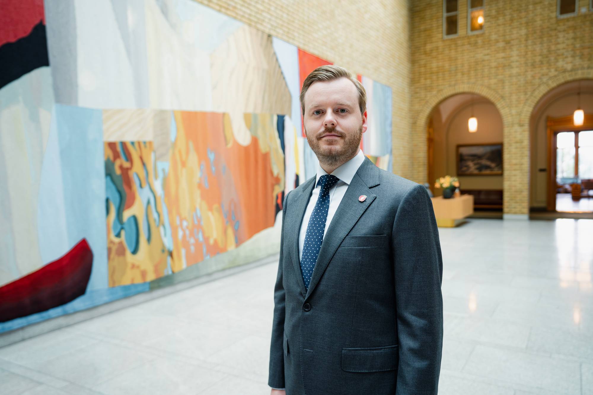 Ein mann står inne i vandrehallen på Stortinget. På veggen er eit stort veggteppe. Mannen har på seg grå dress, kvit skjorte og slips. Han har brunt hår og kort skjegg. 