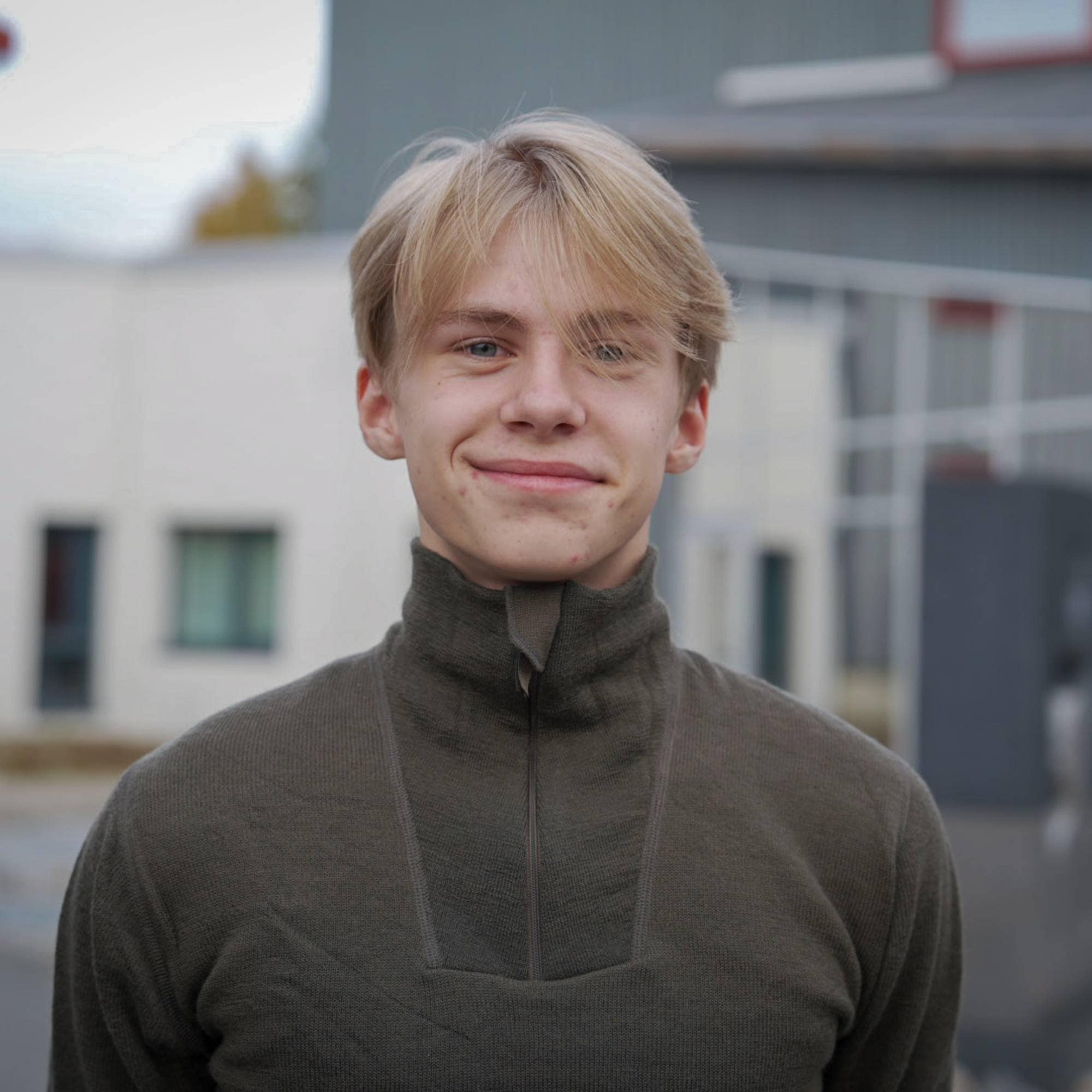 Portrett av en ung mann med blondt hår i en olivenfarget genser foran en bygning. (Bildebeskrivelsen er laget av en KI-tjeneste)