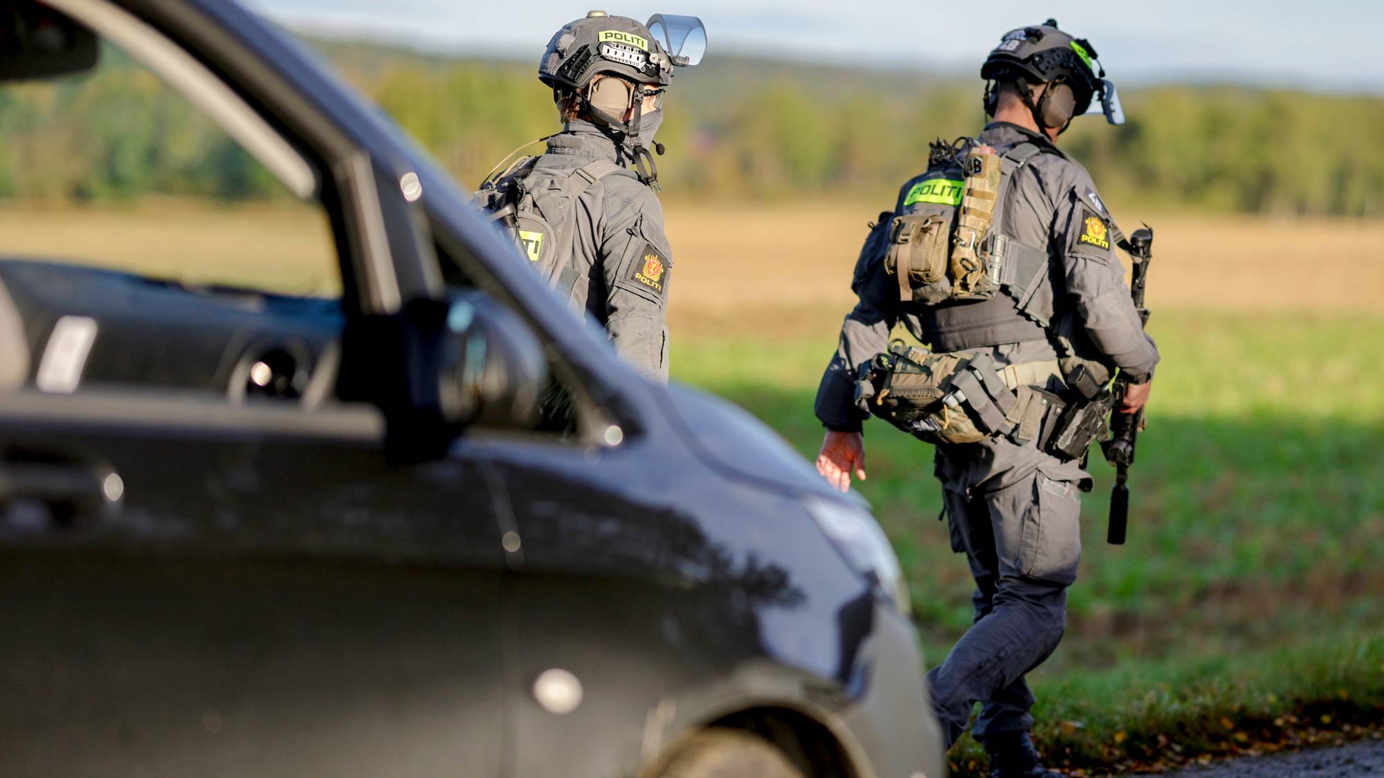 To bevæpnede polititjenestepersoner i uniform går langs en vei ved et jorde. (Bildebeskrivelsen er laget av en KI-tjeneste)