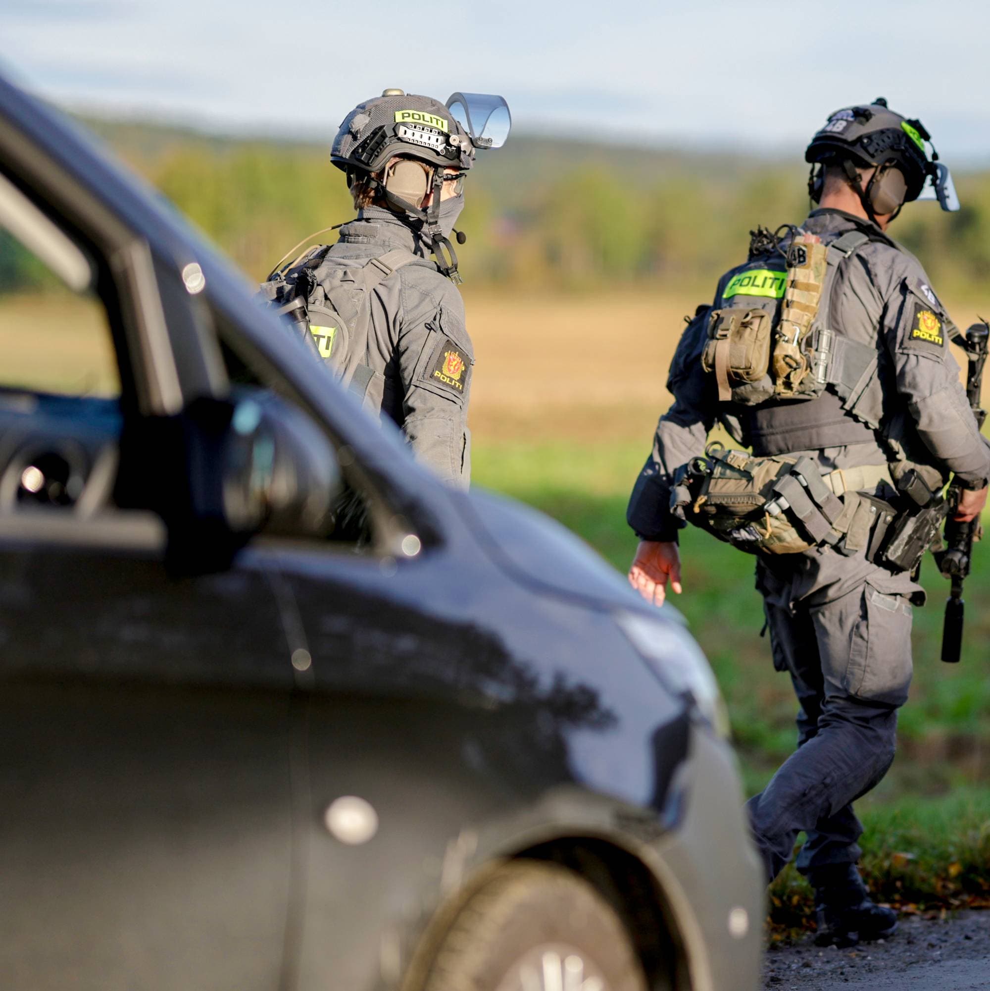 To bevæpnede polititjenestepersoner i uniform går langs en vei ved et jorde. (Bildebeskrivelsen er laget av en KI-tjeneste)