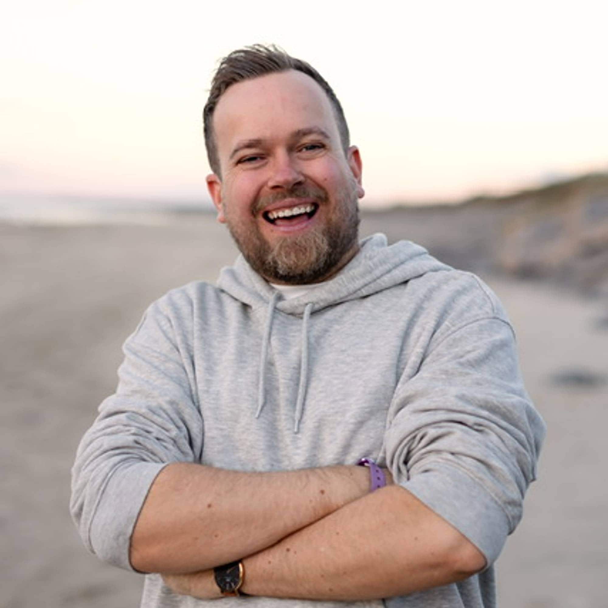 Kjetil Moen smiler med hendene i kryss. Han har en grå genser på seg og står på en strand.