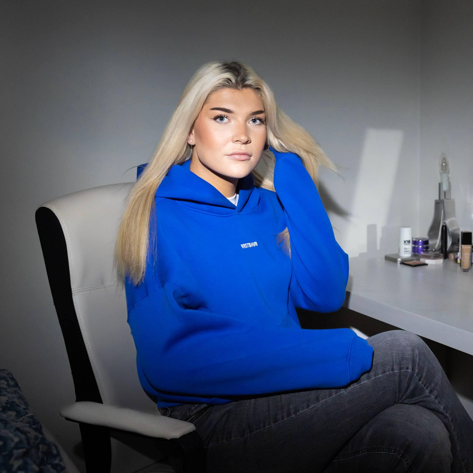 Celine (20) sitter ved sminkebordet på rommet hennes. Hun har på seg en jeans og en blå hettegenser. Hun har langt blondt hår med midtskill. Hun løfter litt av håret sitt med venstre hånd mens hun ser i kamera med et mykt blikk.