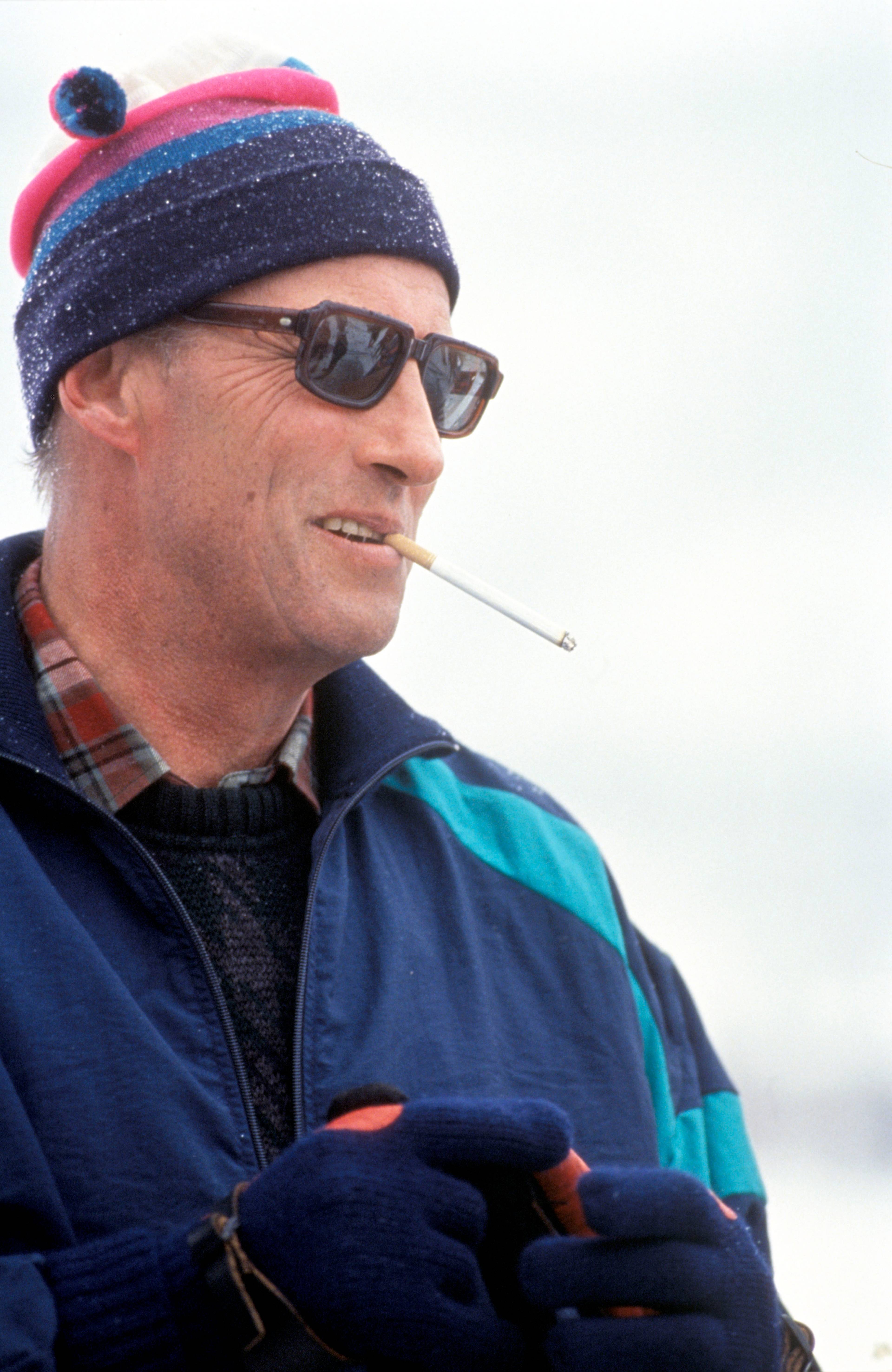 Ein mann med hatt, solbriller og sigarett i munnen. Han er iført jakke og hanskar. Bakgrunnen er uskarp og lys. (Bildebeskrivelsen er laget av ein KI-teneste)