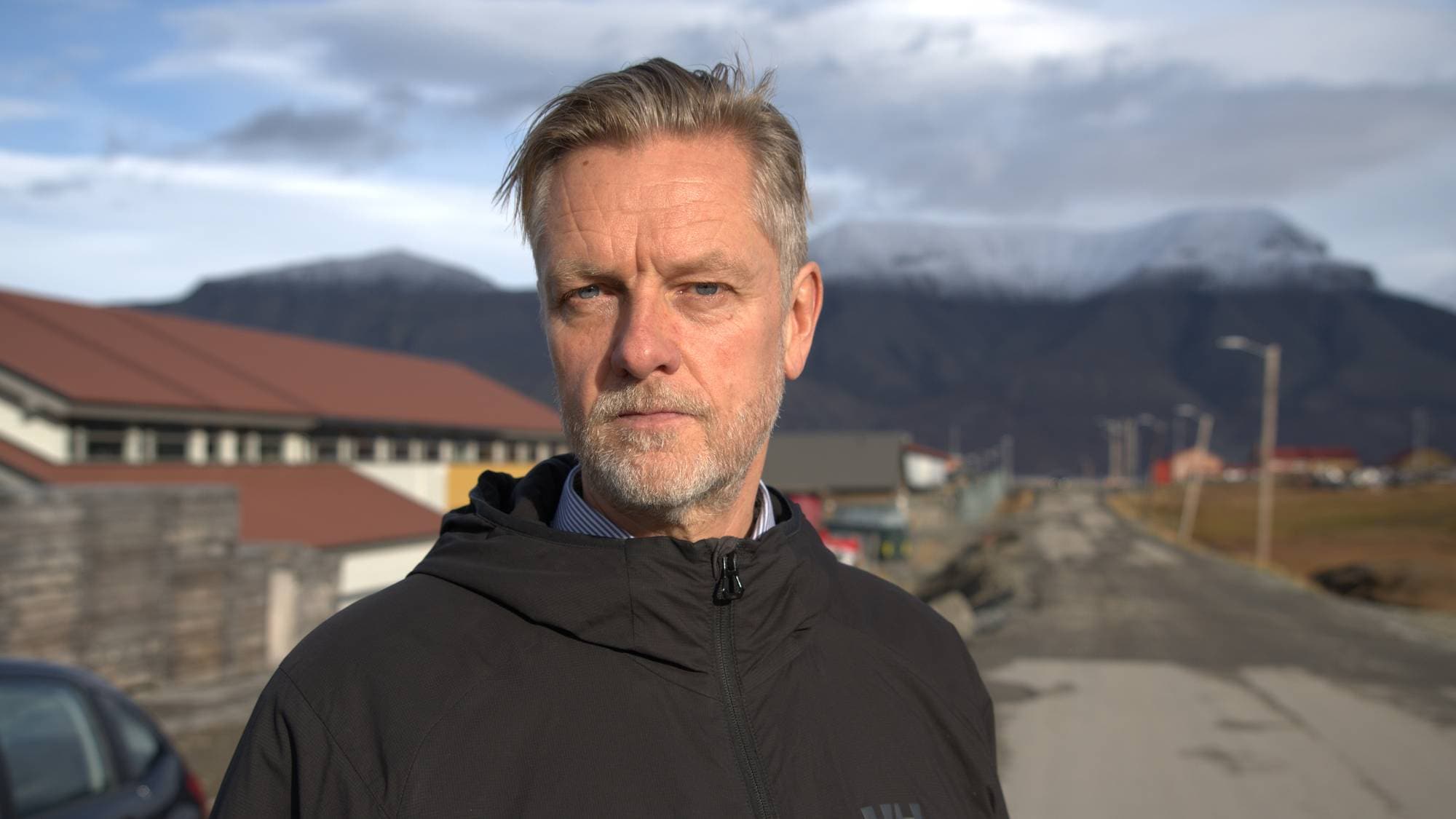 En mann ser i kamera. Fjell og bygninger i bakgrunn. 