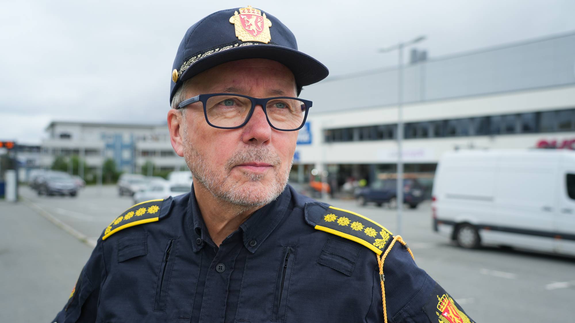 En politimann i uniform og med politilue utendørs ved en parkeringsplass. (Bildebeskrivelsen er laget av en KI-tjeneste)