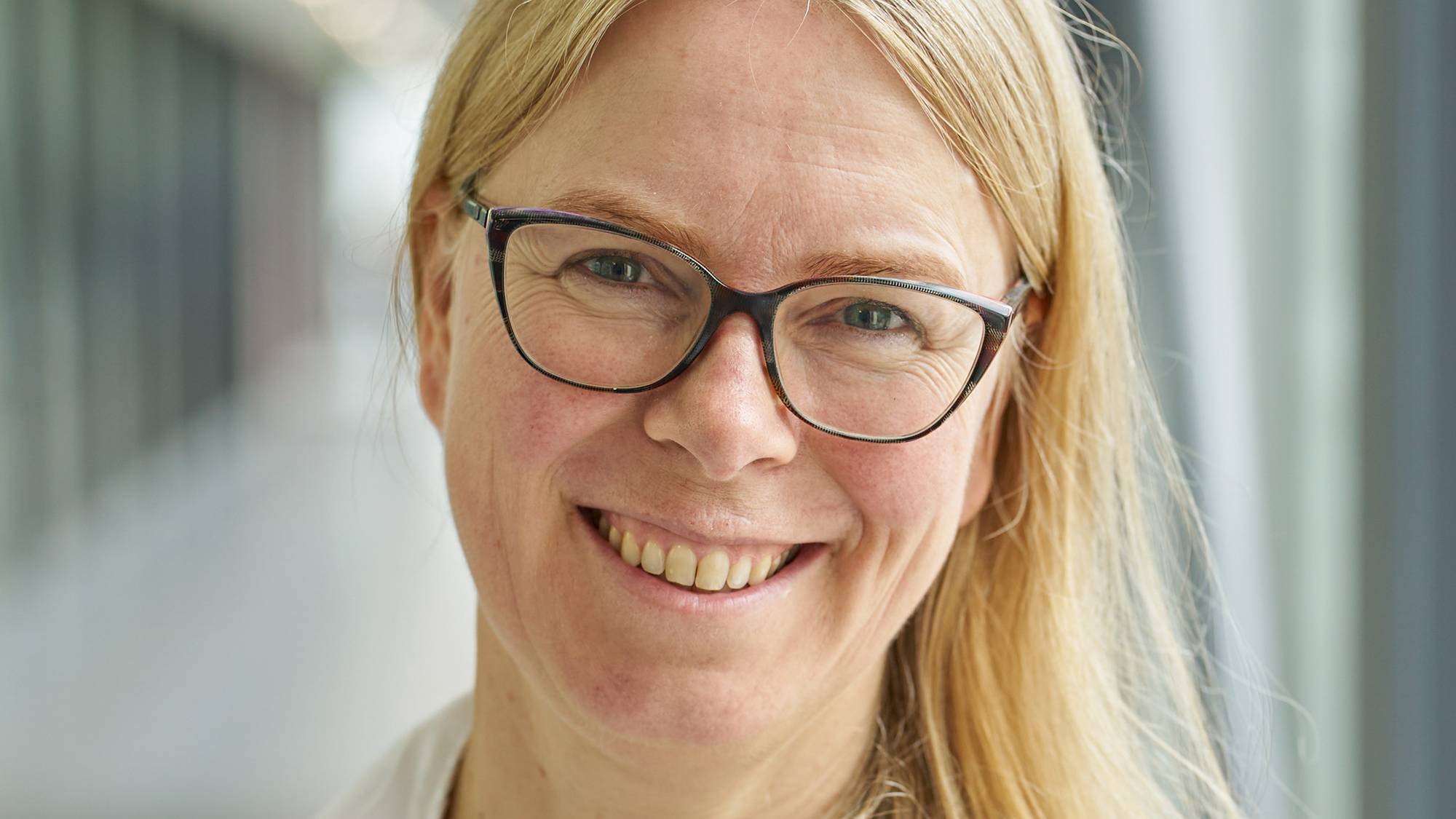 En kvinne står i en gang og smiler, hun er blond og har briller.