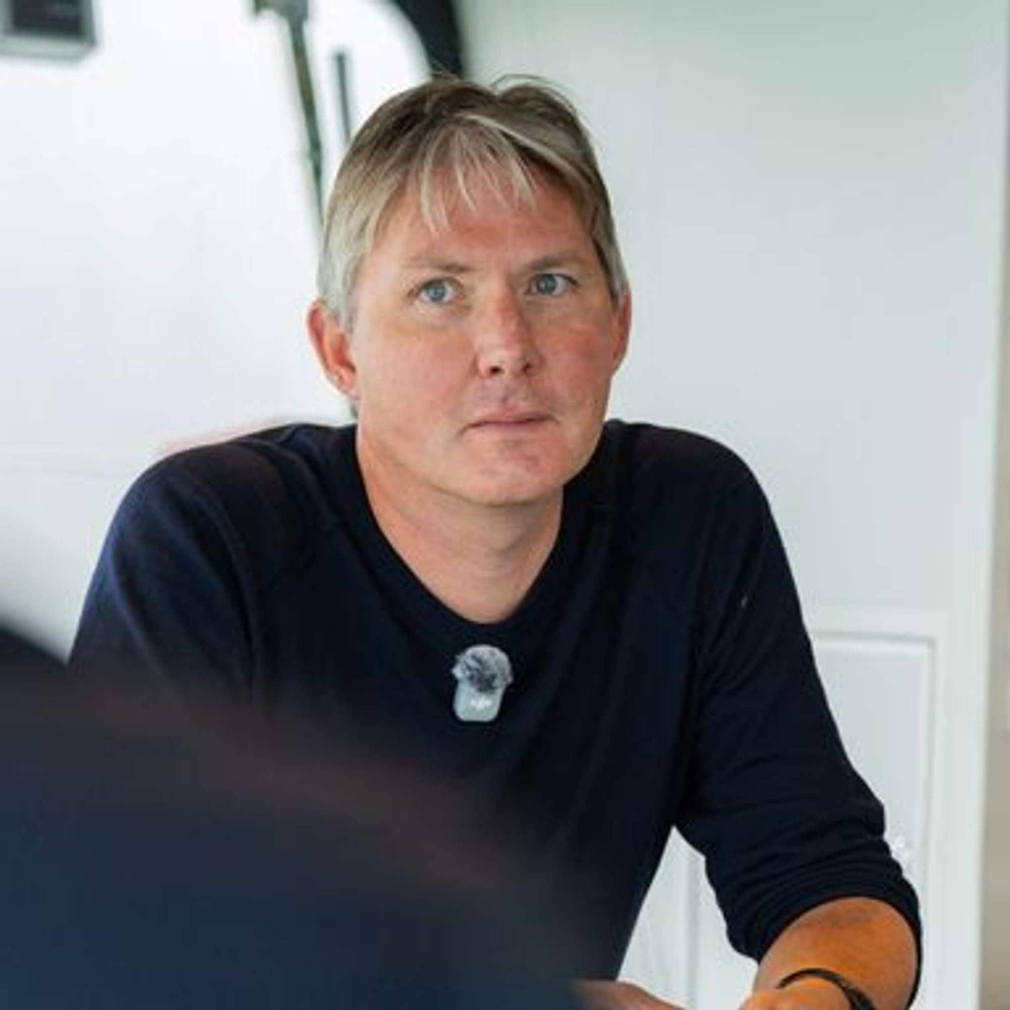 En mann i mørk genser sitter innendørs og ser mot siden under et intervju. 