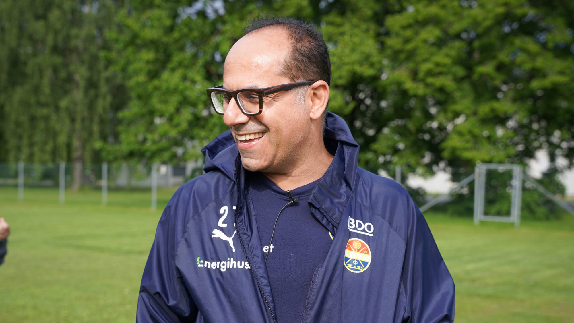 Mann med briller og blå jakke smiler ute på en gressplen. Han har på seg en blå Godset-jakke. I bakgrunnen er det trær og en fotballbane. (Bildebeskrivelsen er laget av en KI-tjeneste)