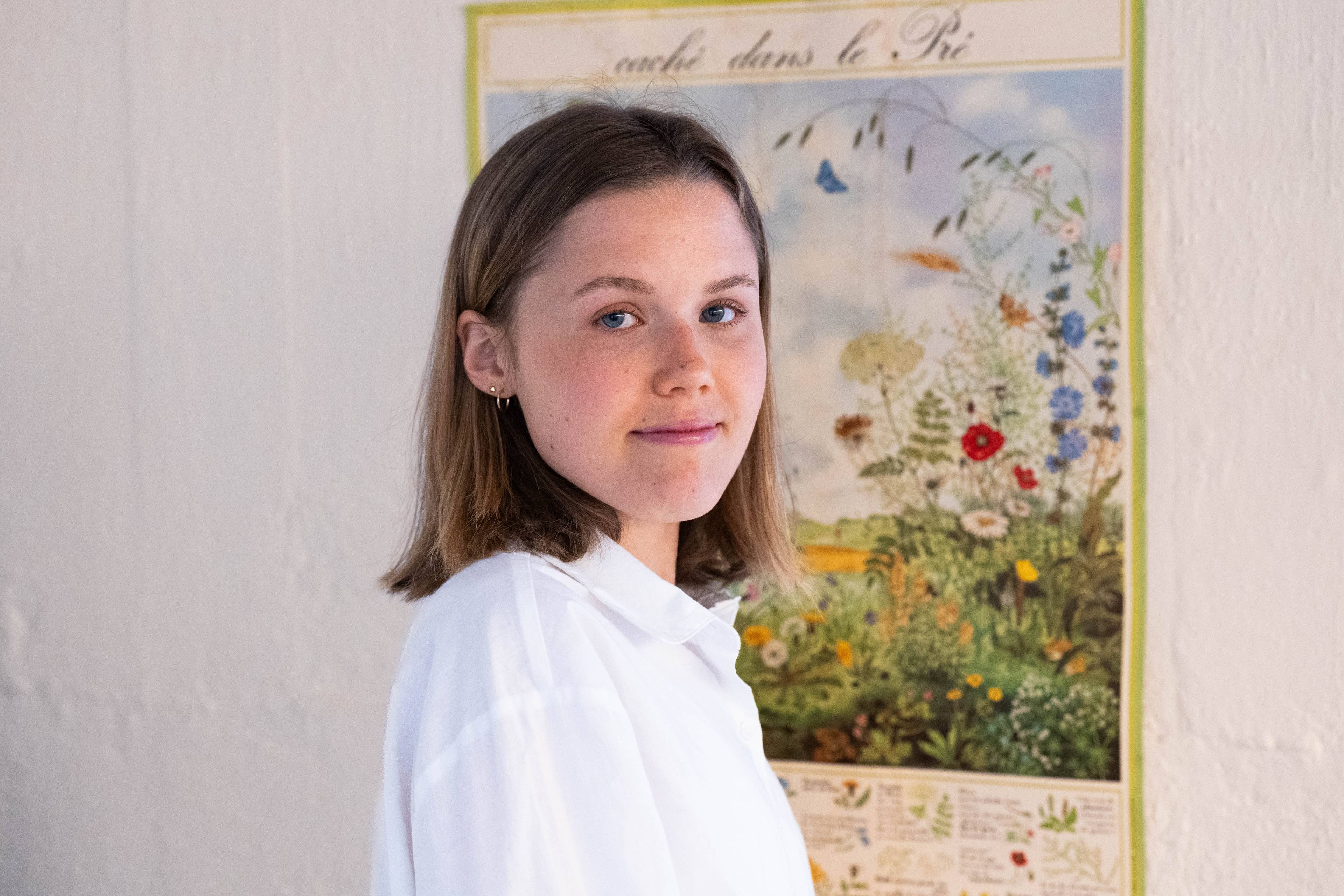 En ung kvinne med kort, brunt hår og blå øyne. Hun står foran en plakat med mange blomster.