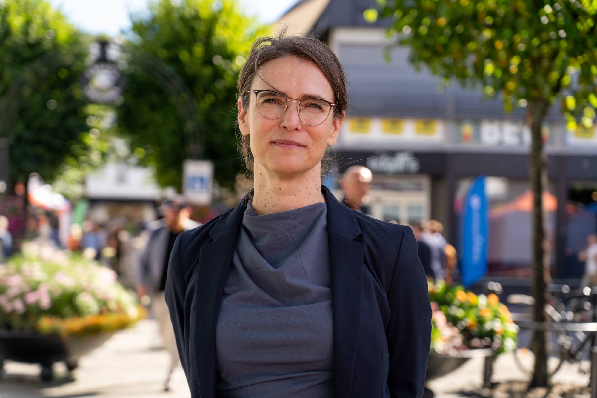 Kvinne med briller og mørk dressjakke står utendørs i en bygate. (Bildebeskrivelsen er laget av en KI-tjeneste)