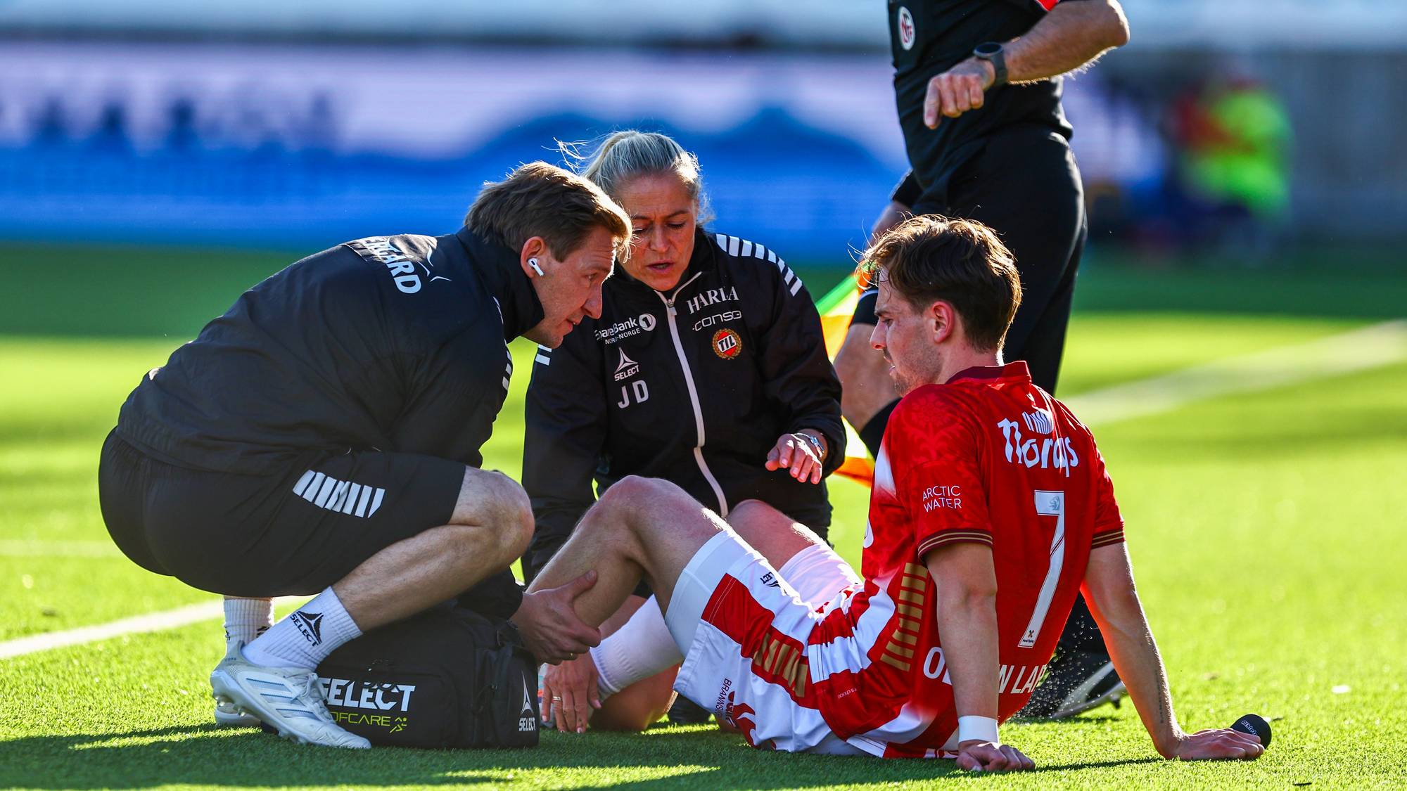 Fotballspiller ligger på bakken under kamp, med medisinsk personell rundt seg. En dommer observerer situasjonen. (Bildebeskrivelsen er laget av en KI-tjeneste)