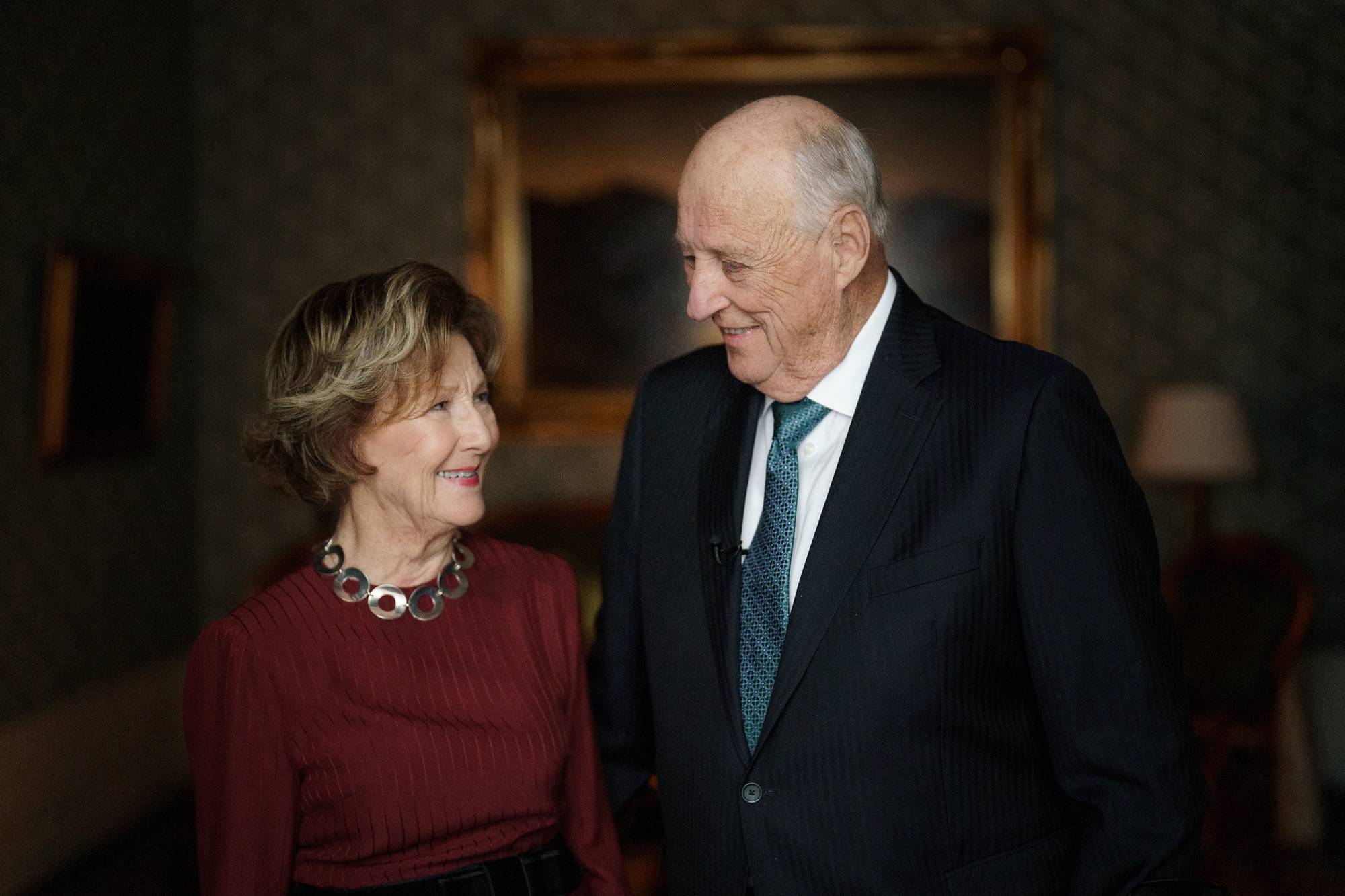 Kong Harald og dronning Sonja står ved siden av hverandre i et rom på Slottet, de ser på hverandre og smiler