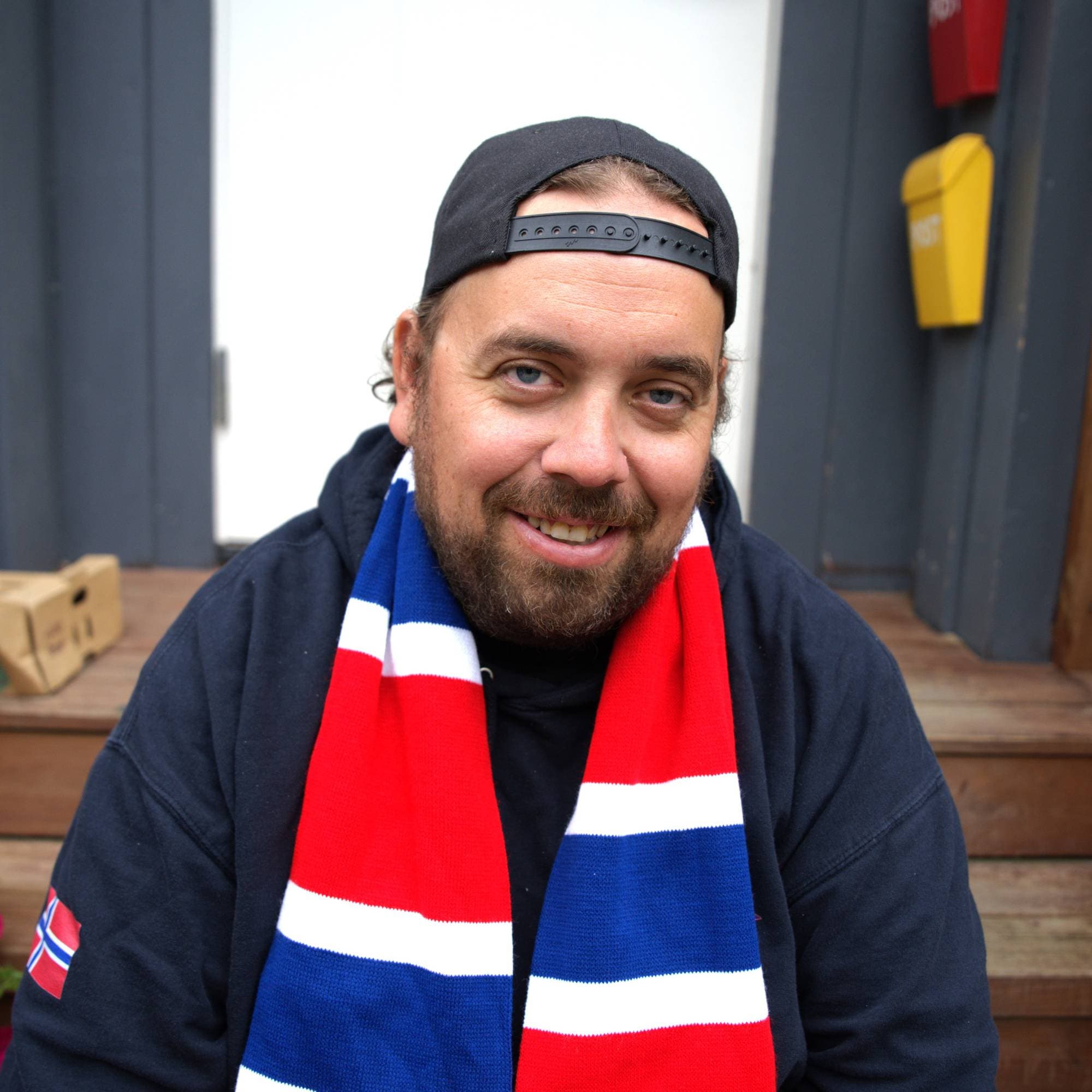 Mann sitter på en trapp foran en hvit dør med hettegenser, caps og supporterskjerf i rødt, hvitt og blått. Han har skjegg og smiler til kamera.