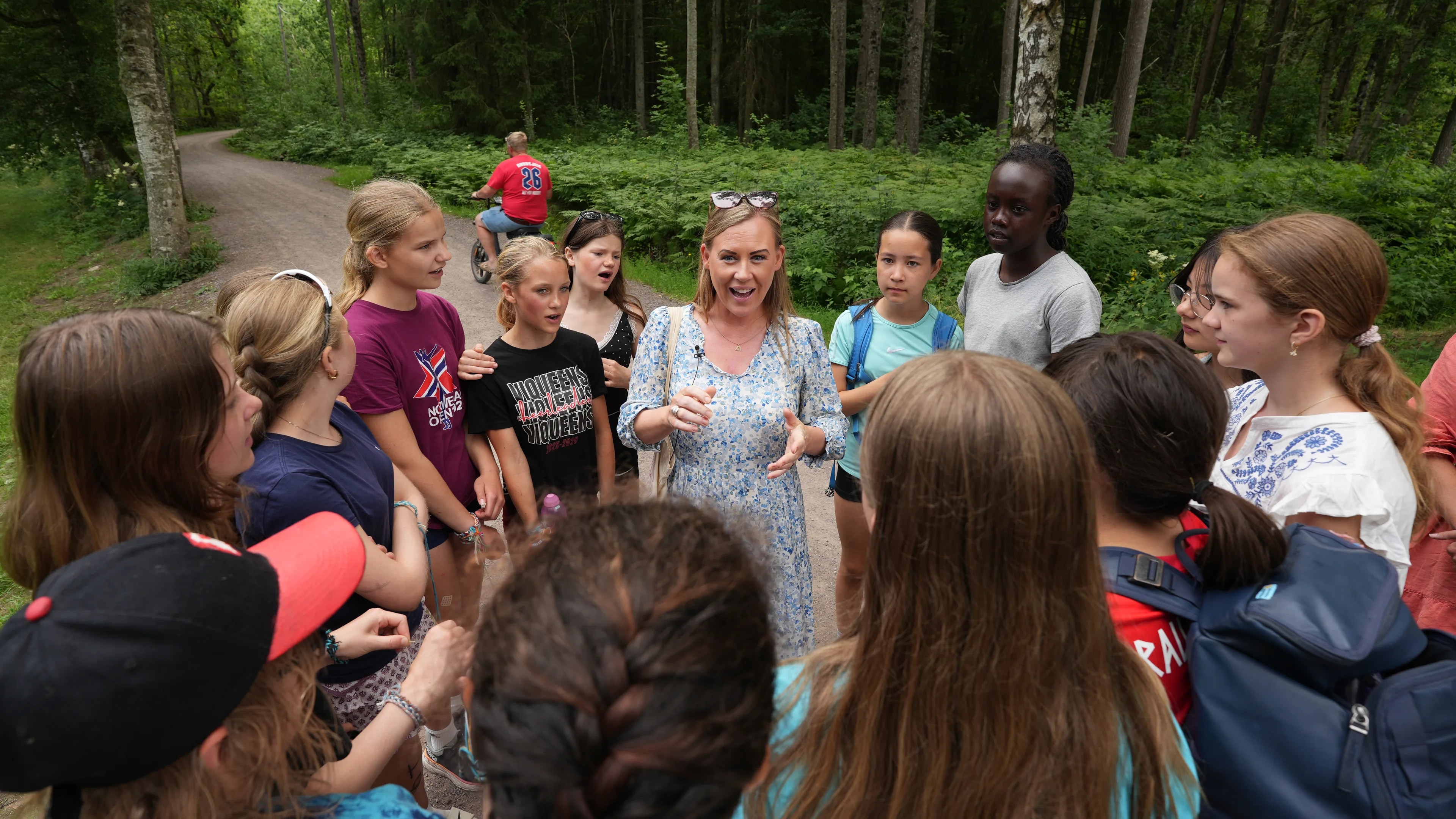 En kvinne snakker til en gruppe barn og ungdommer på en skogsvei. En syklist er synlig i bakgrunnen.