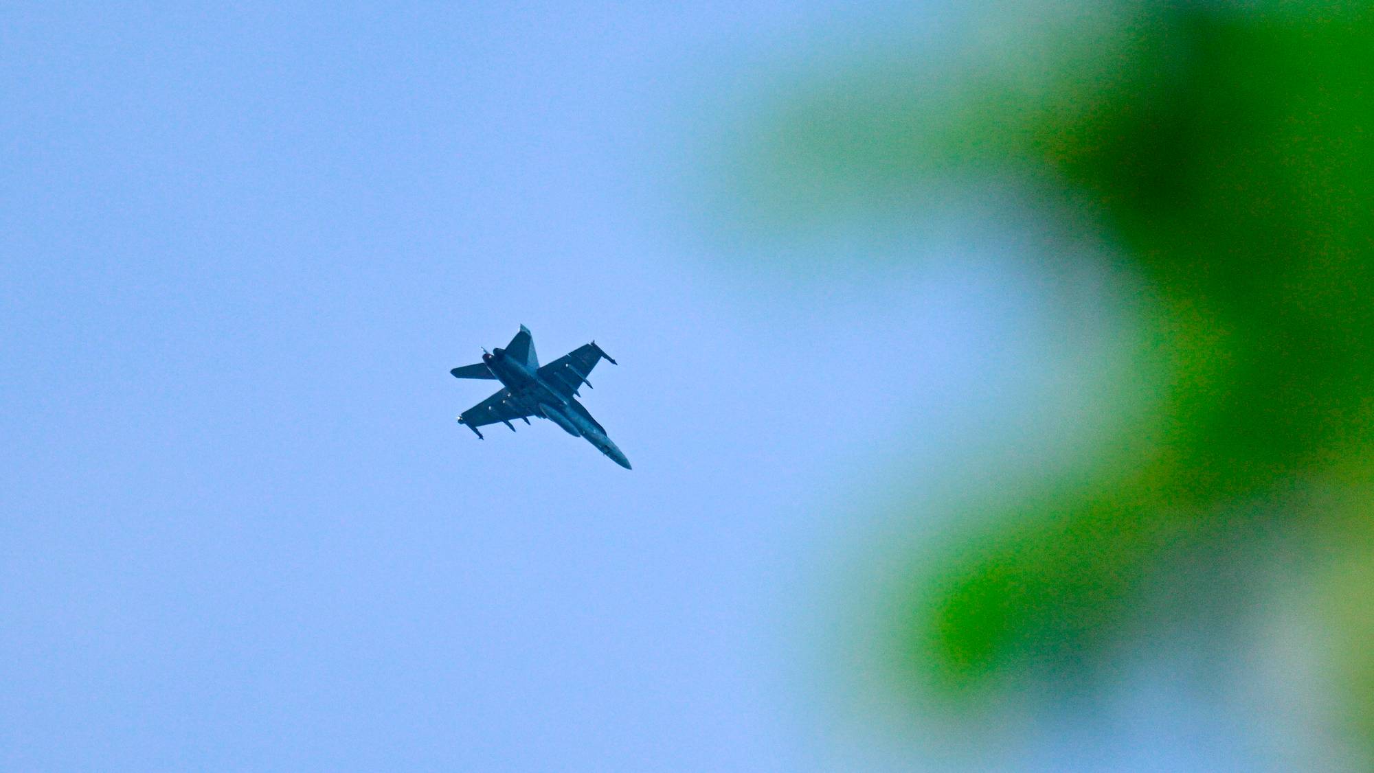 Et kampfly flyr gjennom en klar blå himmel. Et stort, lyst NTB-merke er synlig over flyet. Til høyre er en uskarp grønn masse som kan være et tre. (Bildebeskrivelsen er laget av en KI-tjeneste)