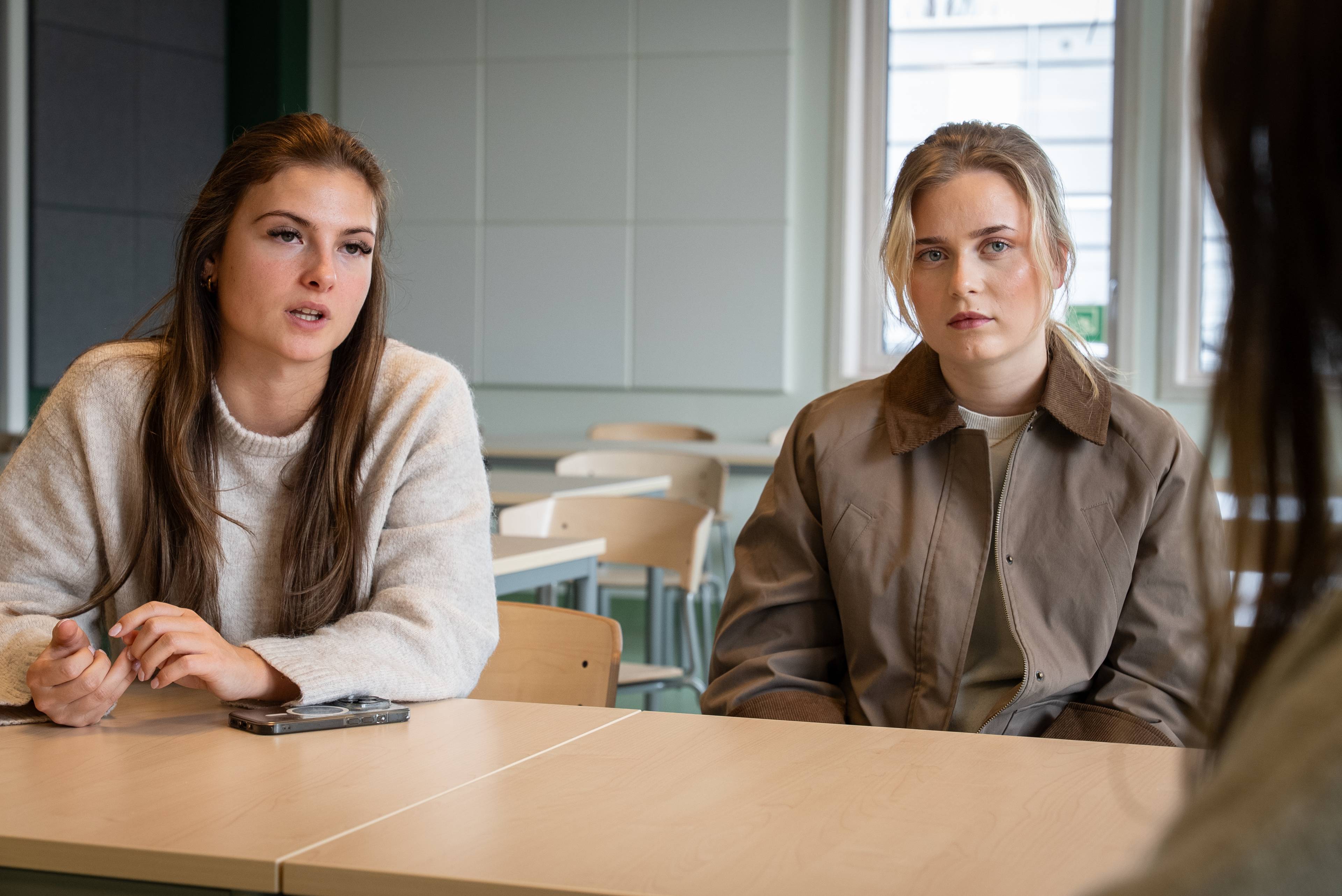 To unge kvinner sitter ved et bord. Den ene kvinnen snakker og gestikulerer. Den andre kvinnen sitter og ser fremover. De sitter i et klasserom. (Bildebeskrivelsen er laget av en KI-tjeneste)