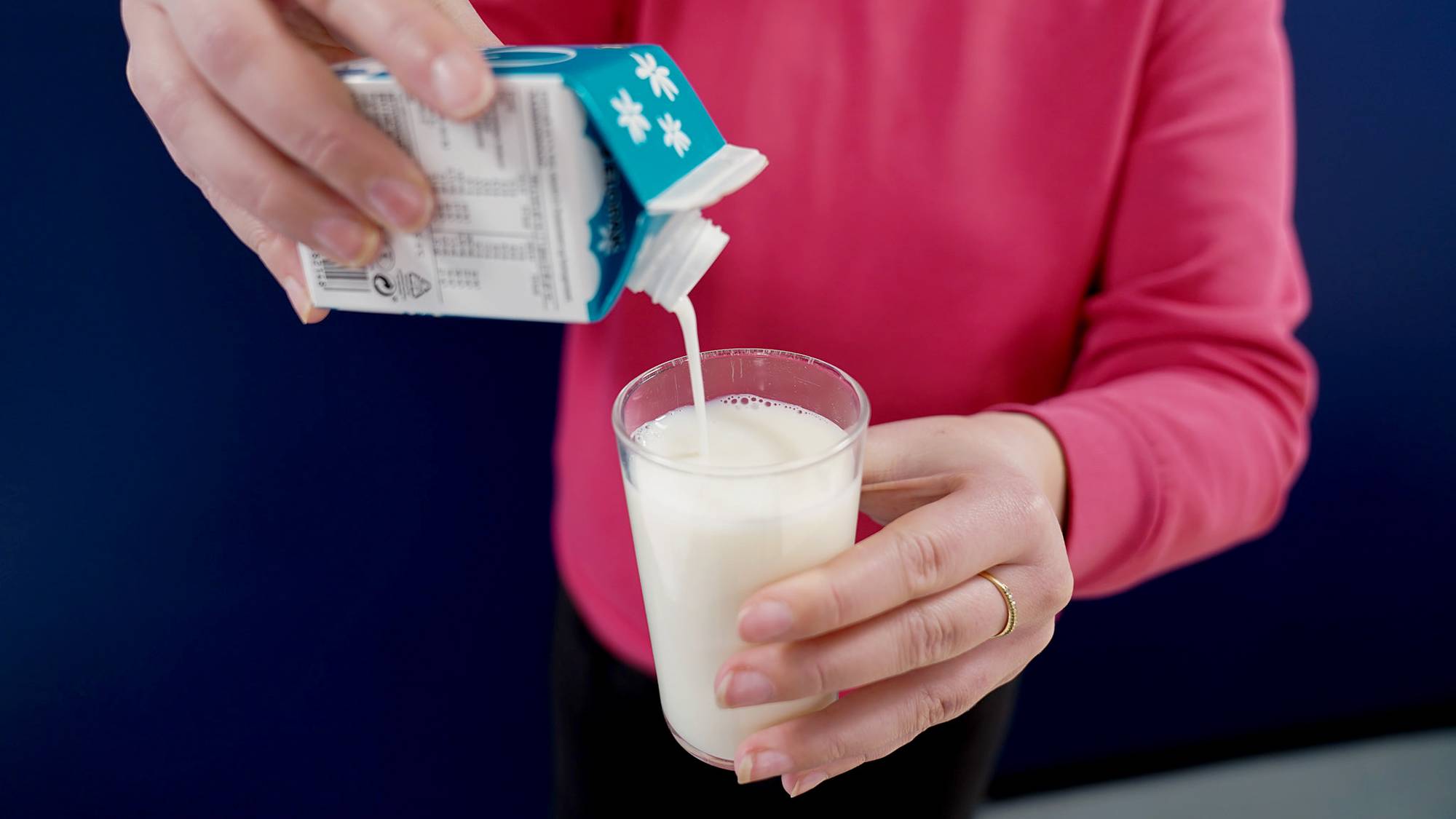 En kvinne heller melk fra en liten grønn kartong i et melkeglass. Hun har på rosa genser og står foran en mørk blå vegg.