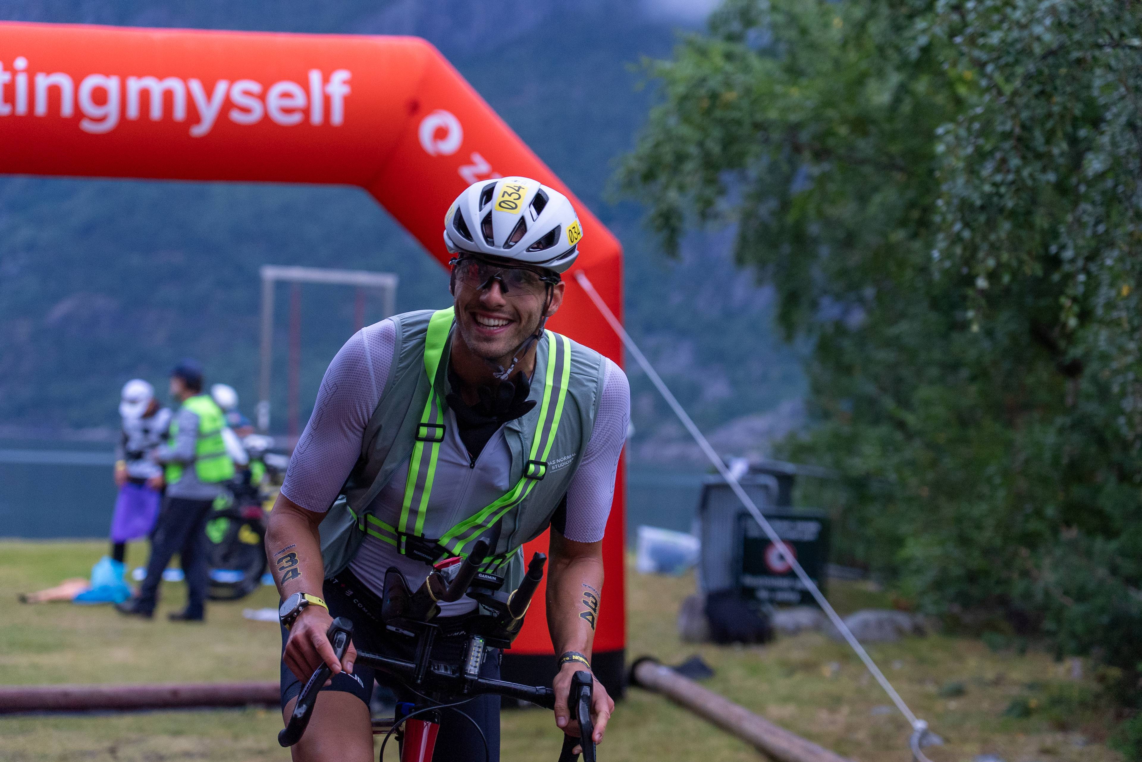 Syklist med hjelm og refleksvest i et løp foran en oppblåsbar portal. Andre deltakere i bakgrunnen. (Bildebeskrivelsen er laget av en KI-tjeneste)