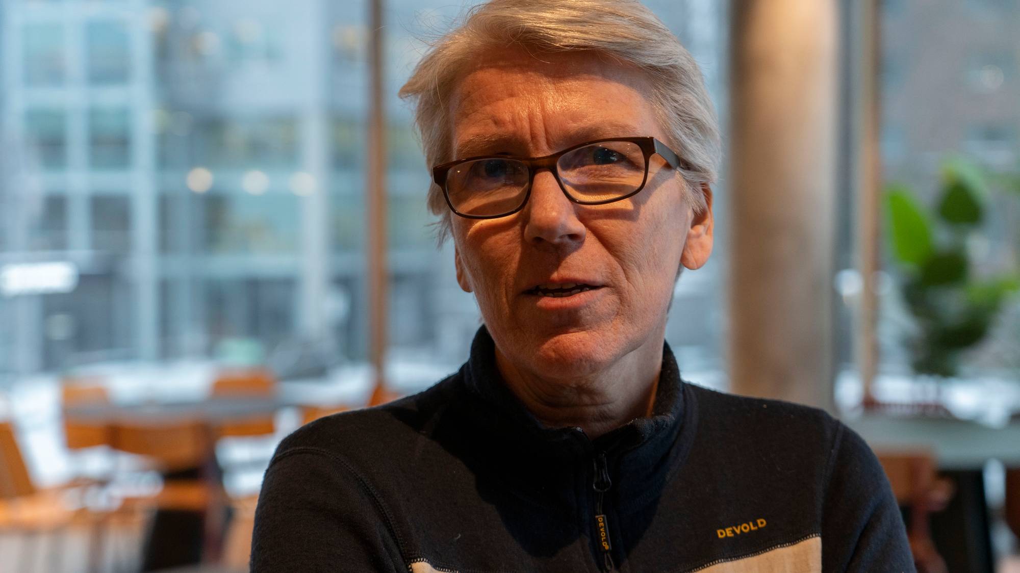 Et nærbilde av en person med grått hår og briller. Personen er kledd i en mørk genser. I bakgrunnen er det et utydelig bilde av et lokale med bord og stoler. (Bildebeskrivelsen er laget av en KI-tjeneste)